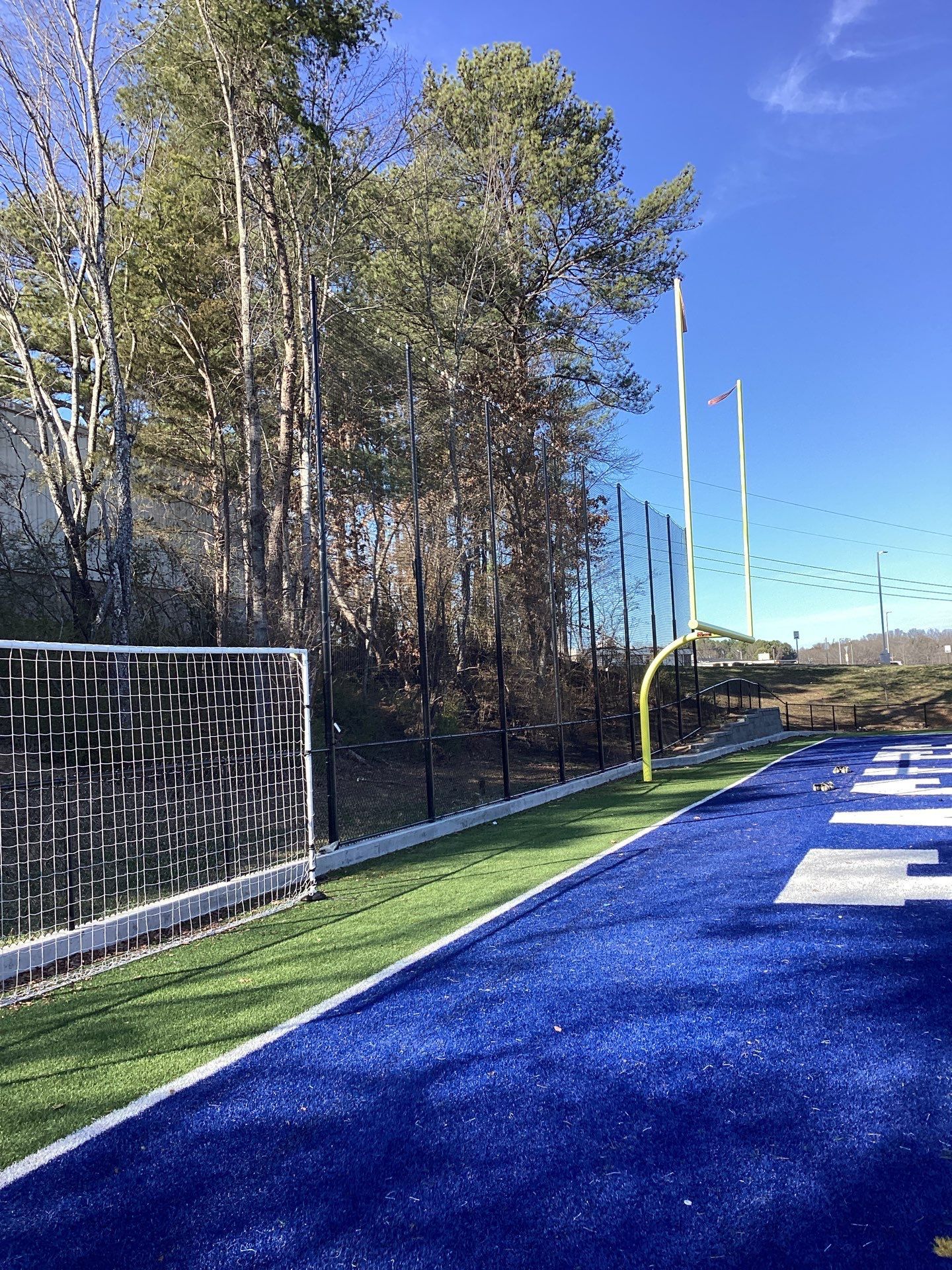 Blue turf football field with goalpost, trees, and fence on a sunny day.