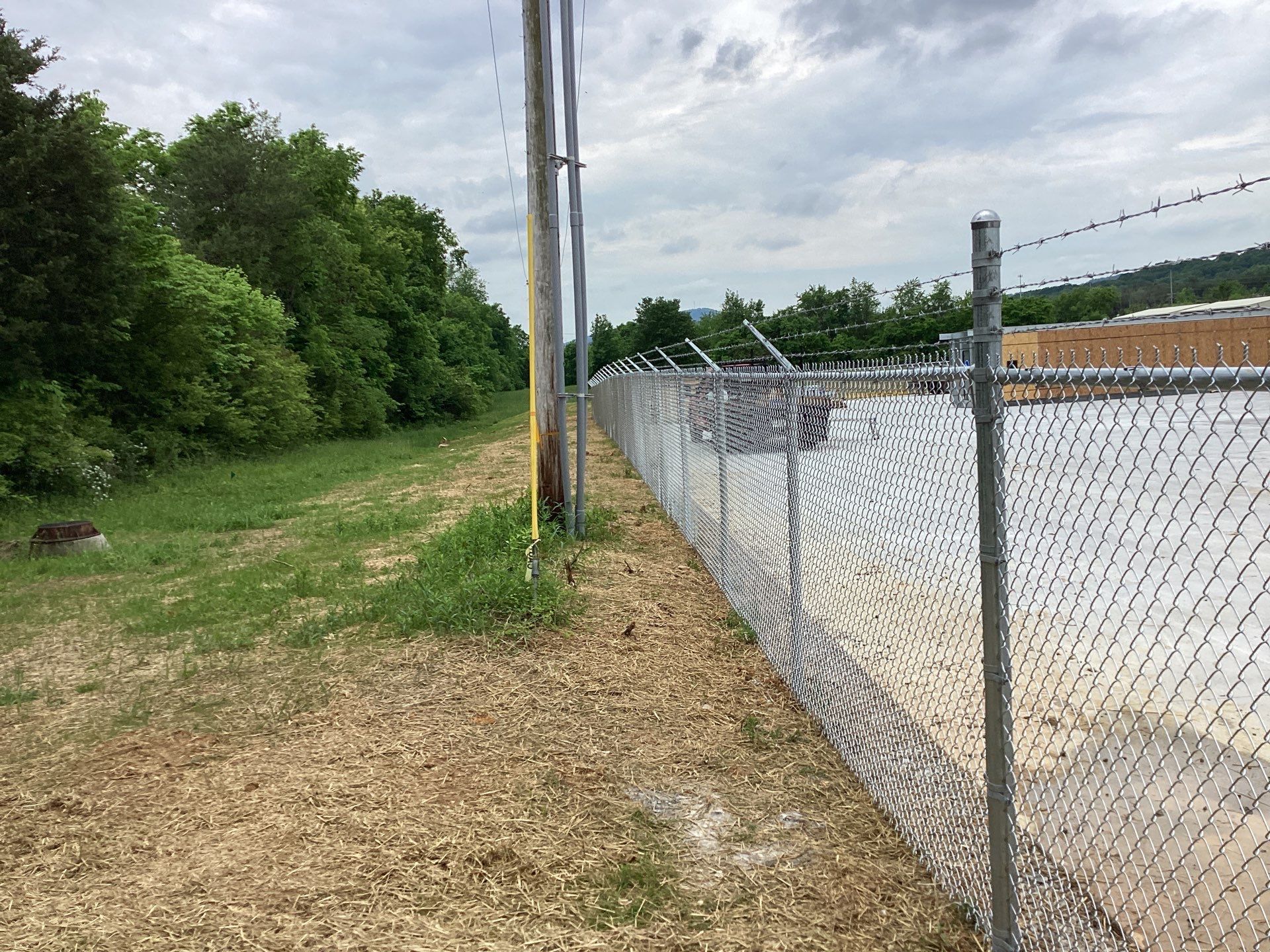 Chain-link fence with barbed wire. Gravel path next to grassy area, bordering a tree line and building. Overcast sky.