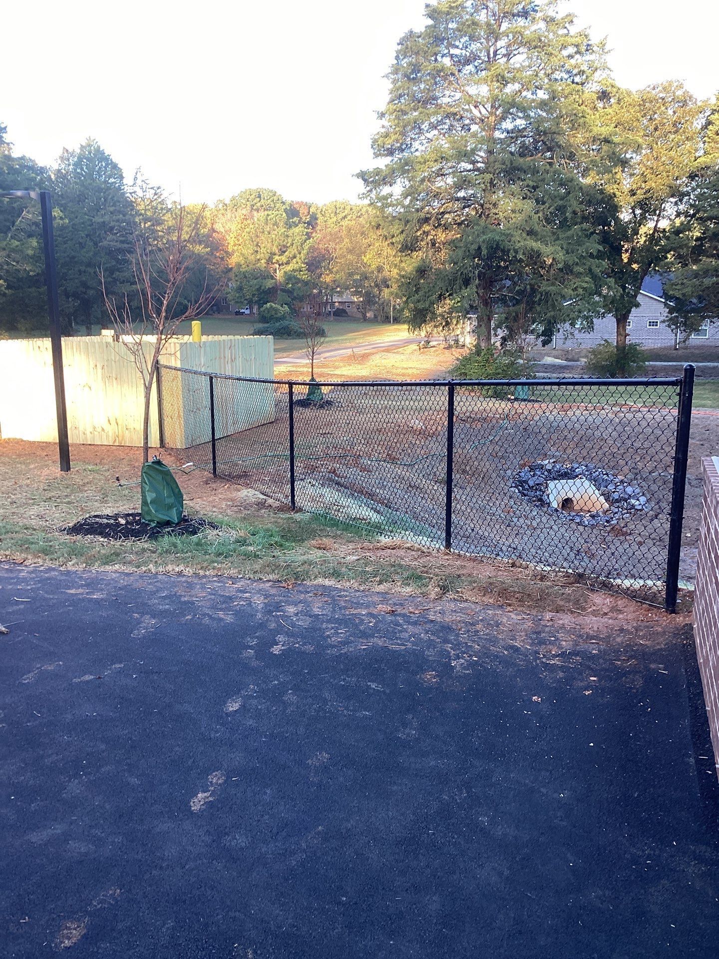 Chain-link fence surrounding a grassy area with trees in the background. Pavement in the foreground.