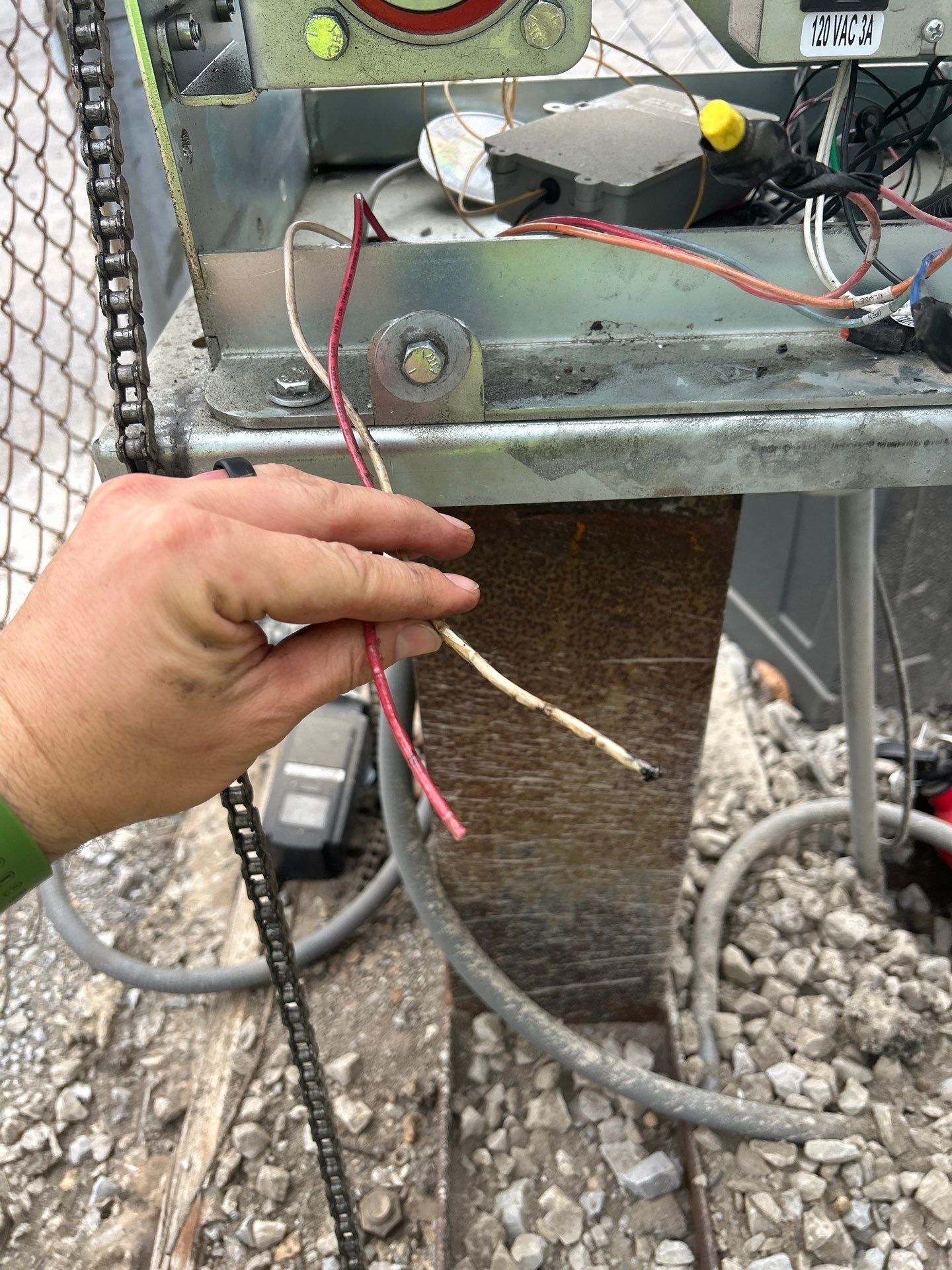 Hand holding cut red and white wires near gate opener machinery; chain, fence and gravel in background.