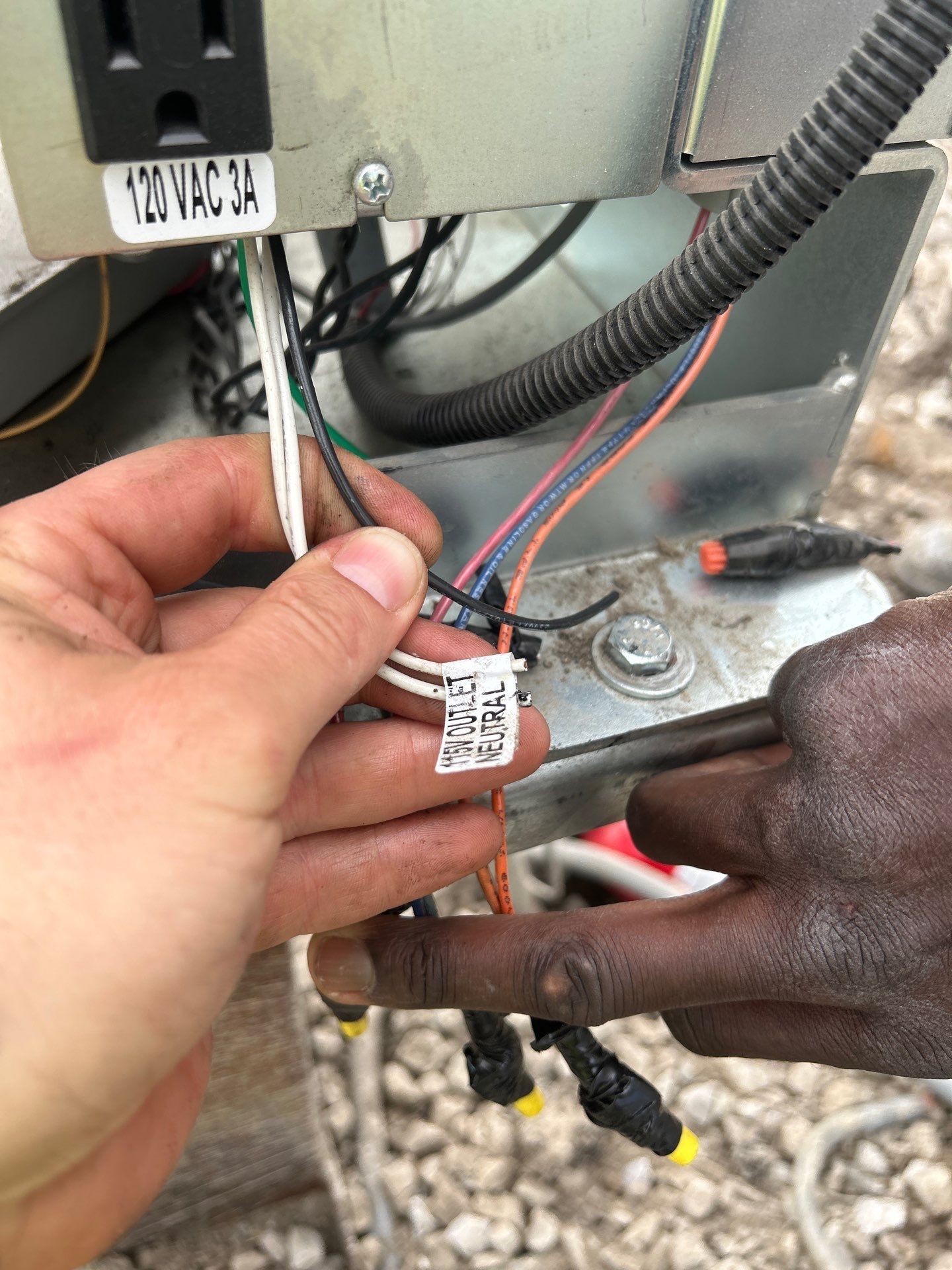 Hands connecting wires on a trailer, near an electrical outlet. Wires are black, white, and orange, on a metal surface.