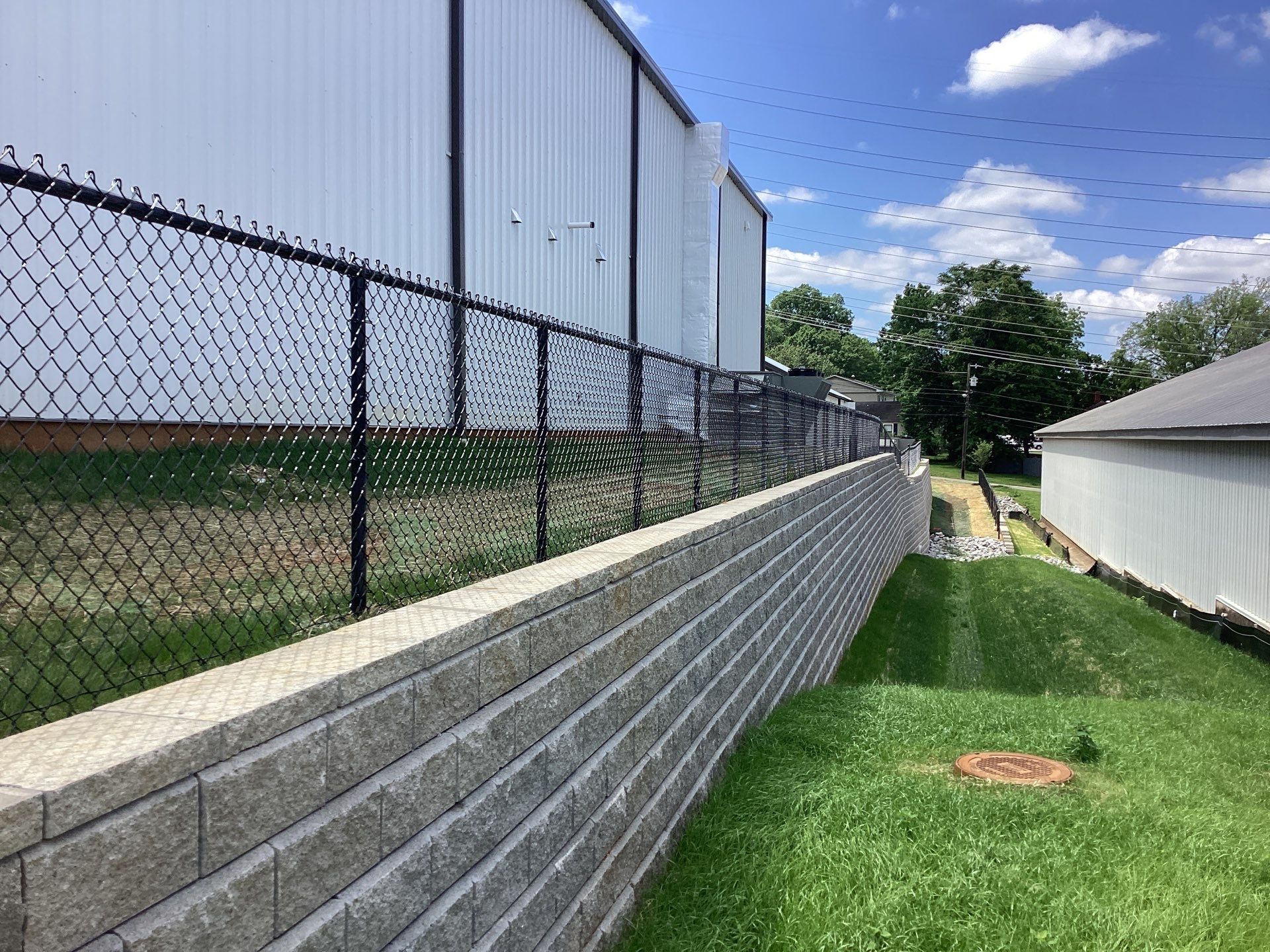 Black chain link fence atop a retaining wall, beside a building with a corrugated metal facade, under a blue sky.