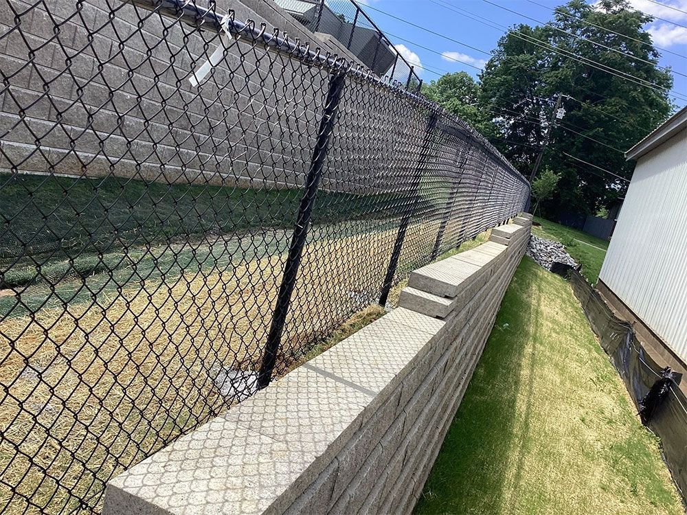 Black chain-link fence atop a retaining wall, along a grassy area, next to a building on a sunny day.