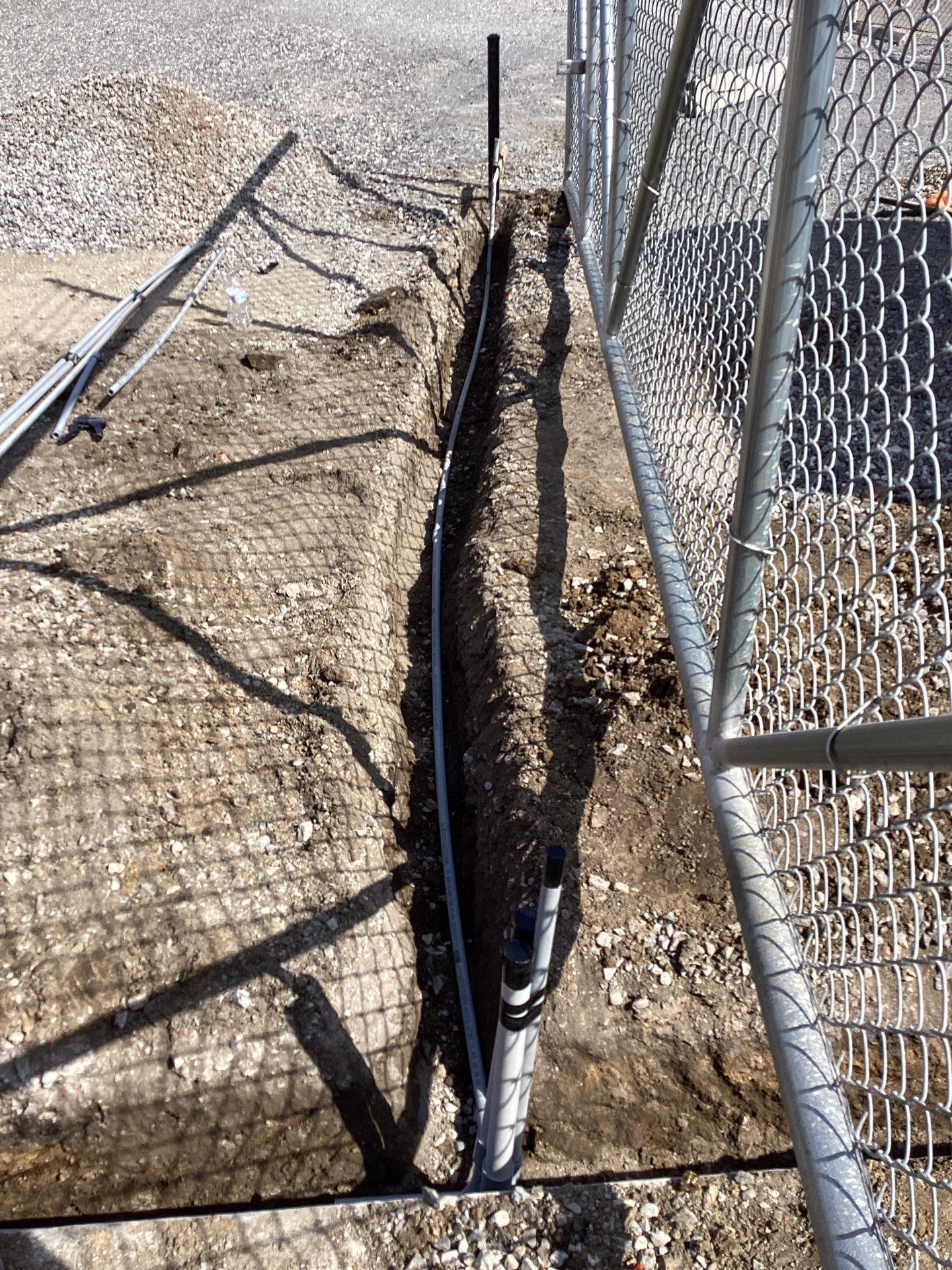 Trench dug next to a chain-link fence, containing black pipes running underground. Sunlight casts shadows.