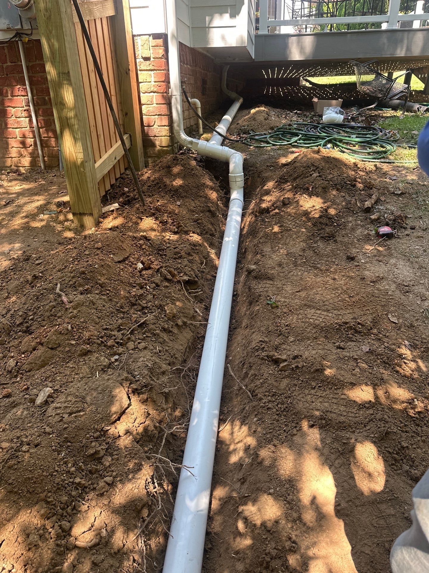 White PVC pipe in a trench, connected to a downspout under a deck. Soil is visible.