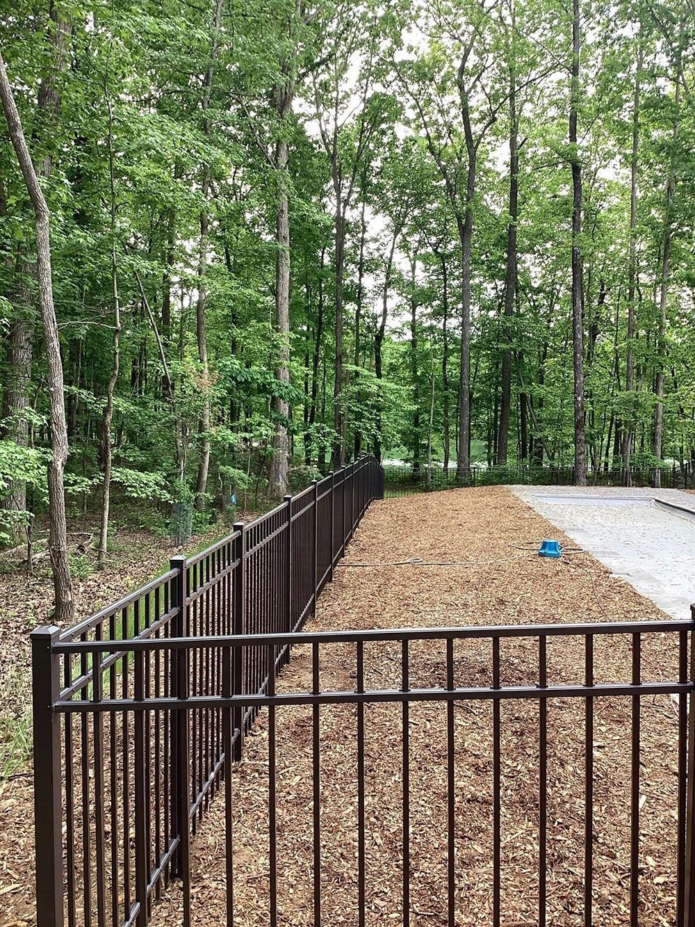 Brown metal fence bordering mulch area with trees in background.