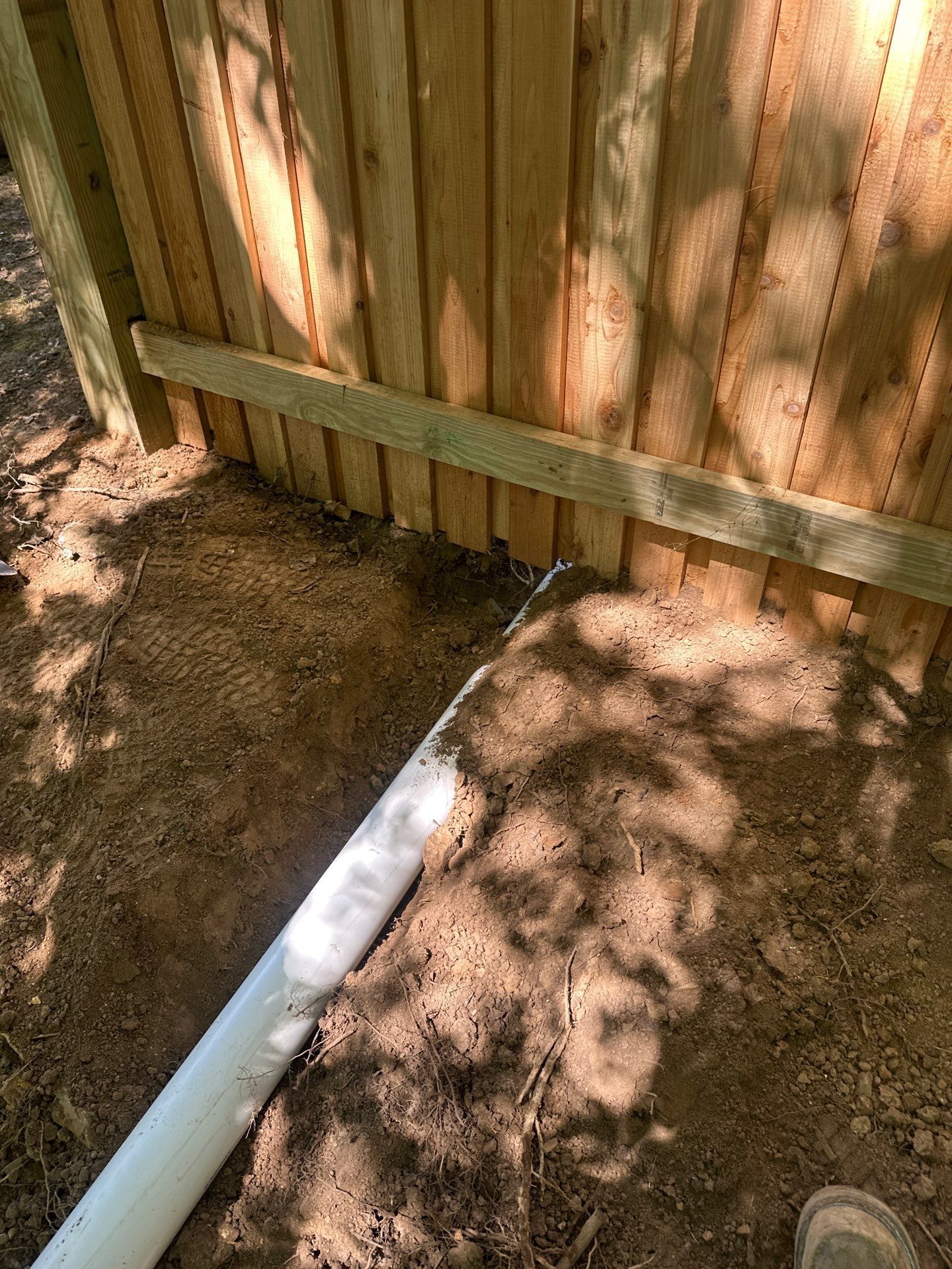 Wooden fence with a horizontal board at the base, and a white pipe lying in the dirt alongside it.