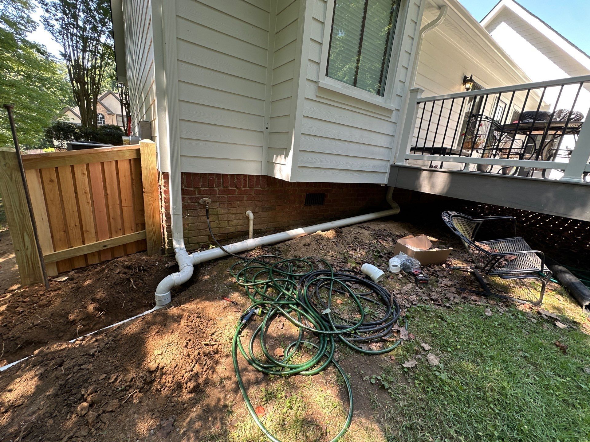Exterior of a house with gutter downspout. A white pipe extends to a disturbed yard area, with a fence and a deck visible.