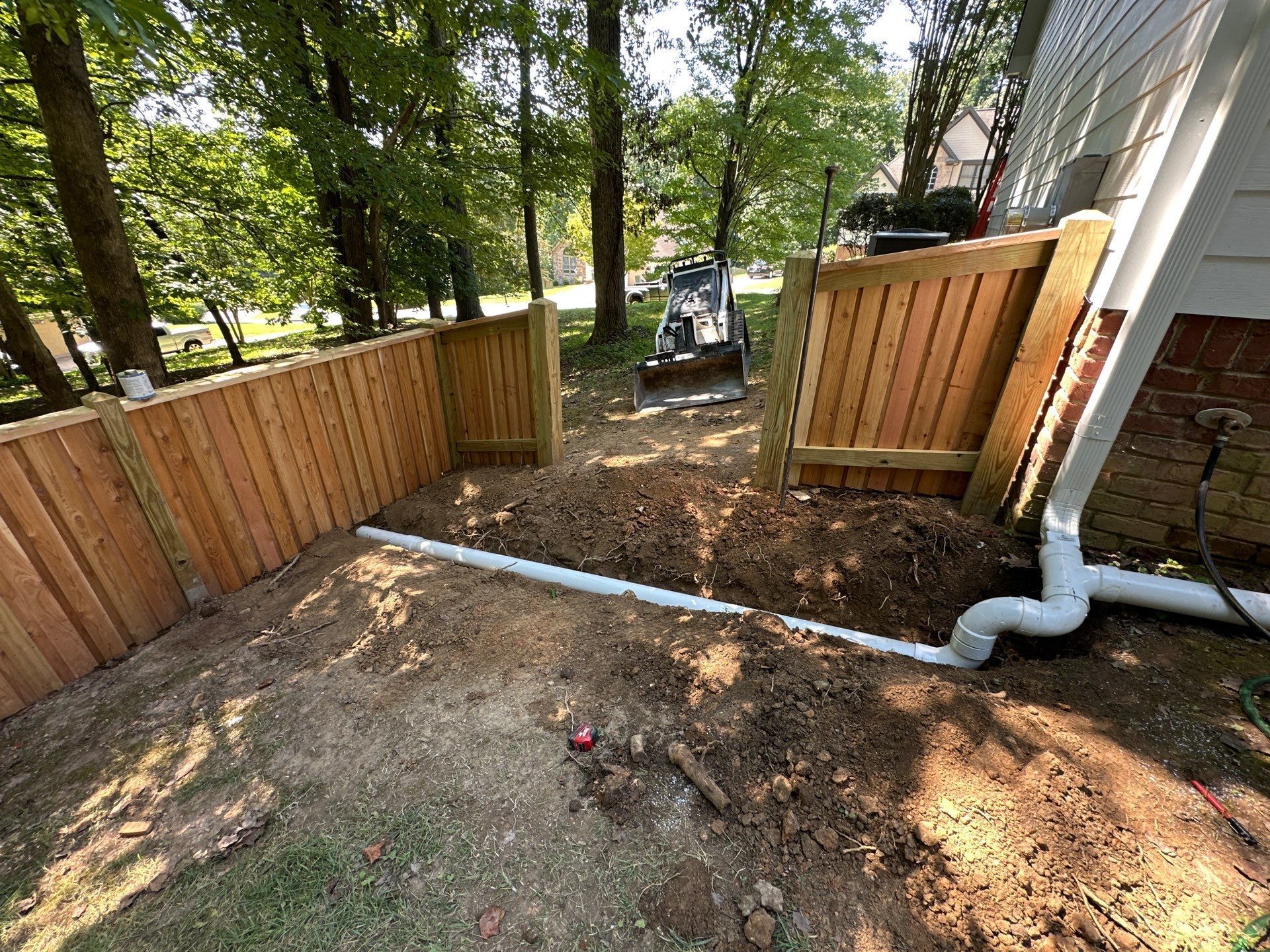 A drainage pipe installation near a fence and house, with a small excavator in the background.