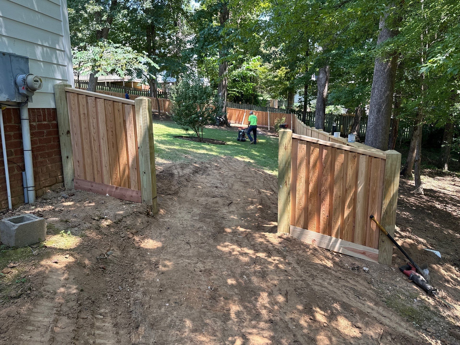 Wooden fence gate on a graded dirt path leading to a grassy yard with a person in the background.