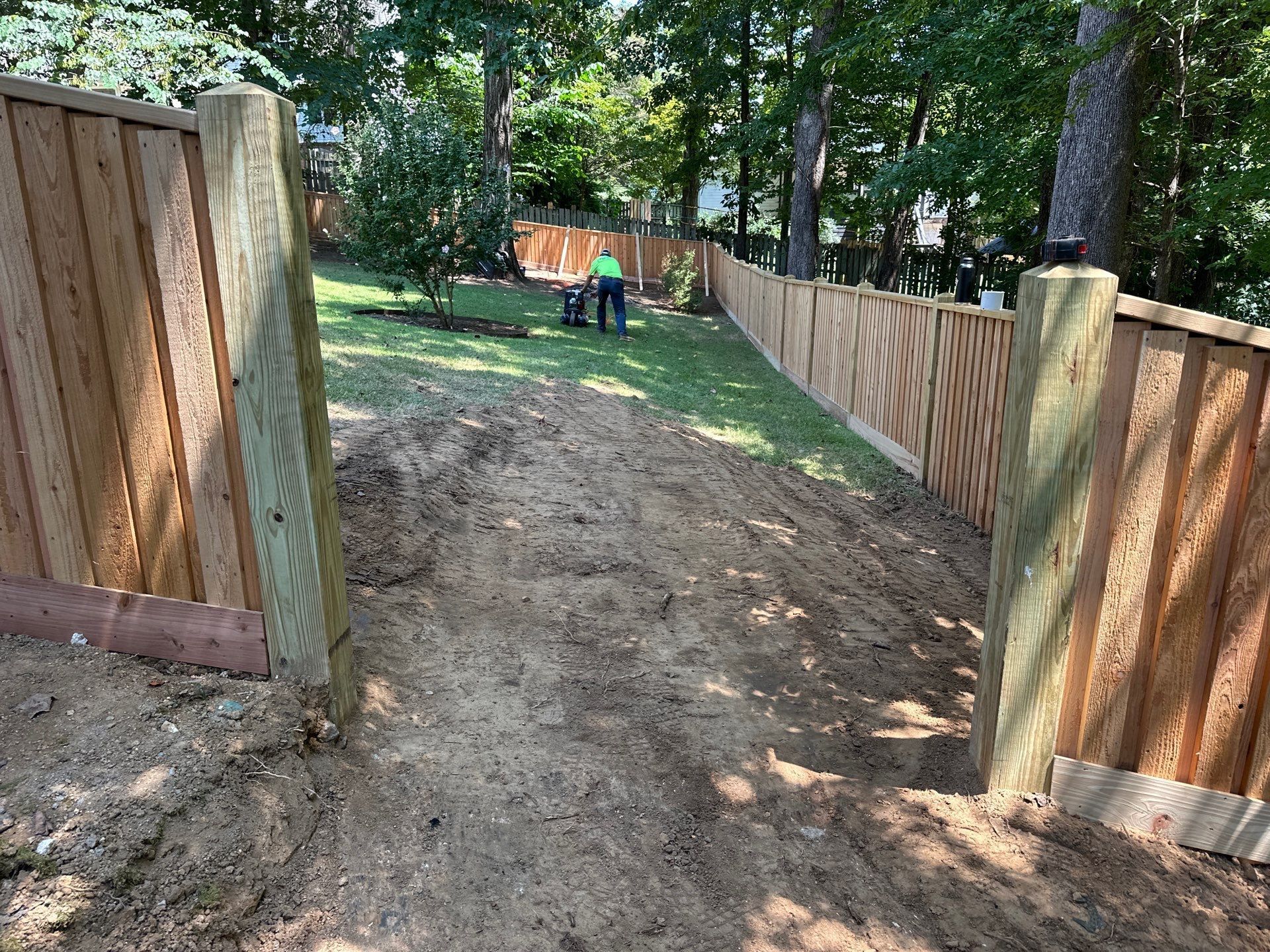 A dirt path leads between two sections of a wooden fence; a person is visible mowing grass in the distance.