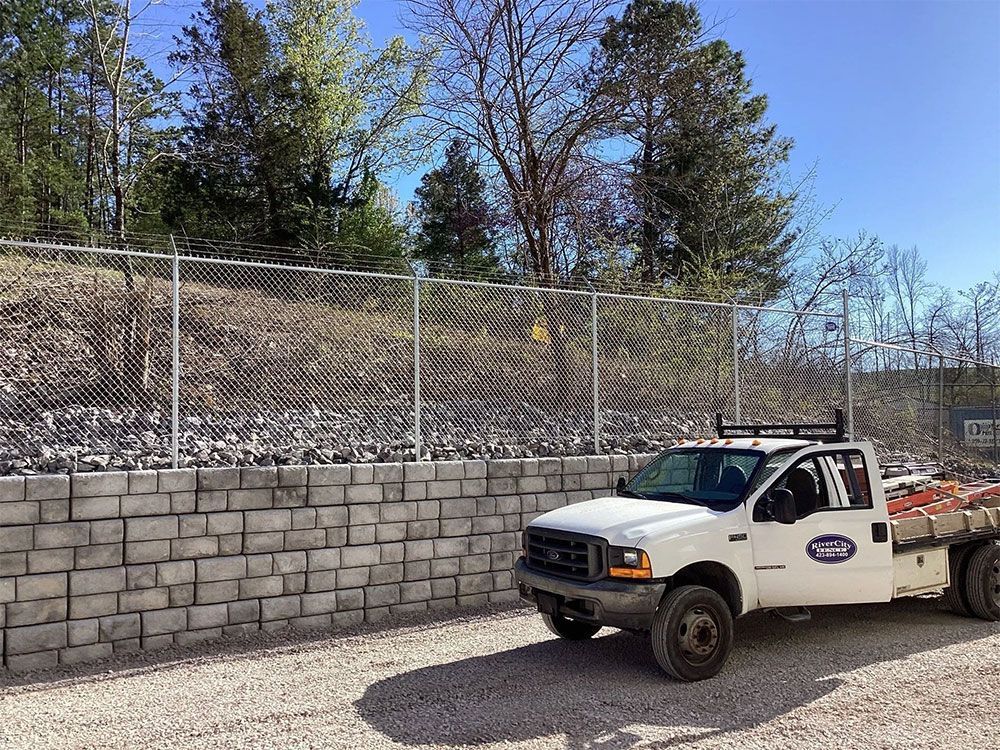 White work truck parked next to a gray stone retaining wall with a chain-link fence on top; trees and a blue sky in the background.