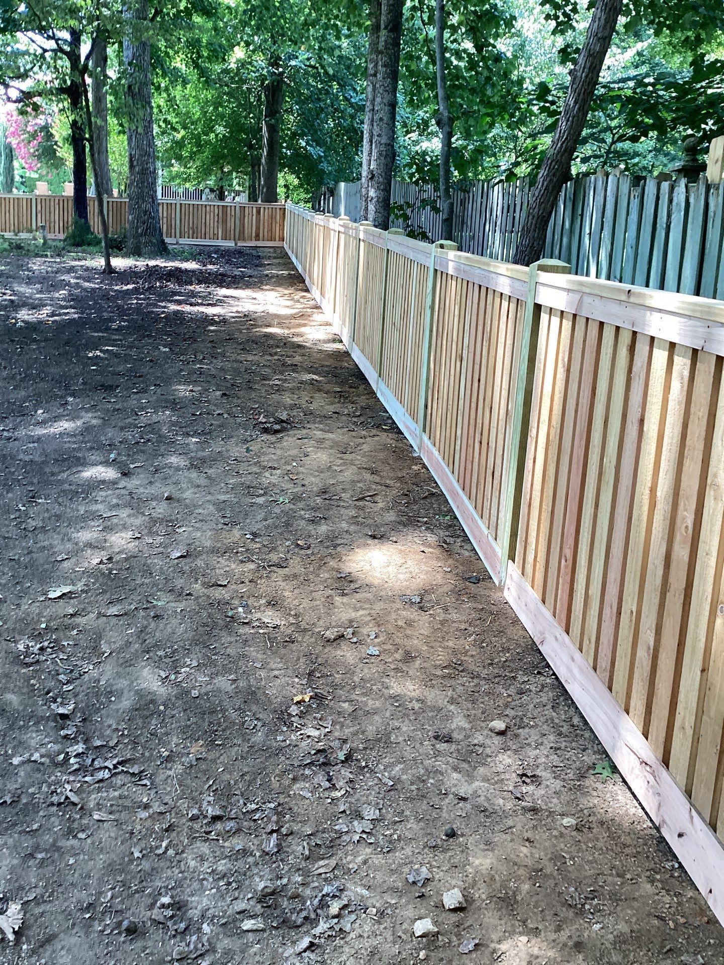 Wooden fence in a yard with dirt ground and trees in the background.