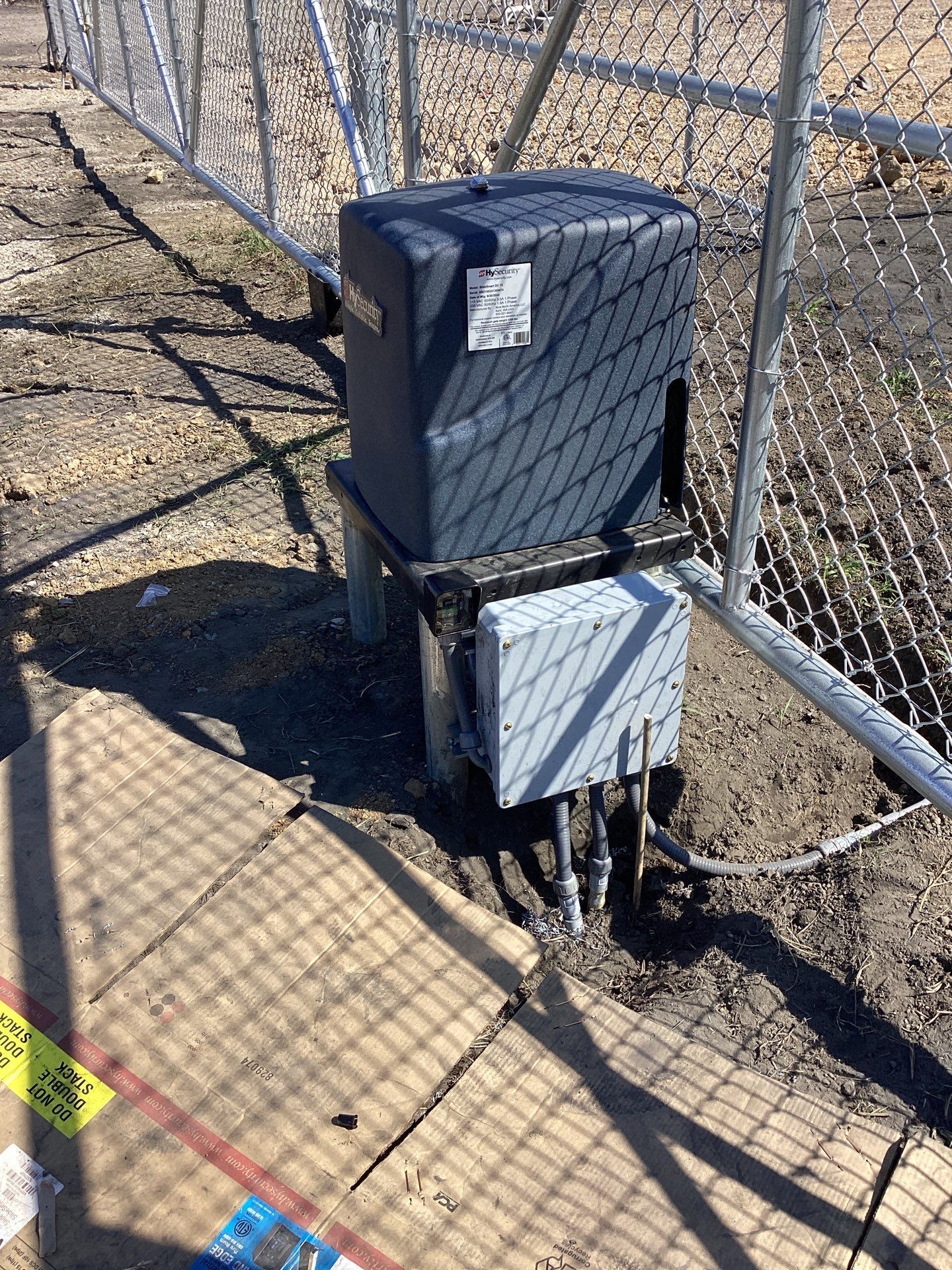 Gate opener system attached to a chain-link fence. Gray box on a metal stand, next to a larger dark box.