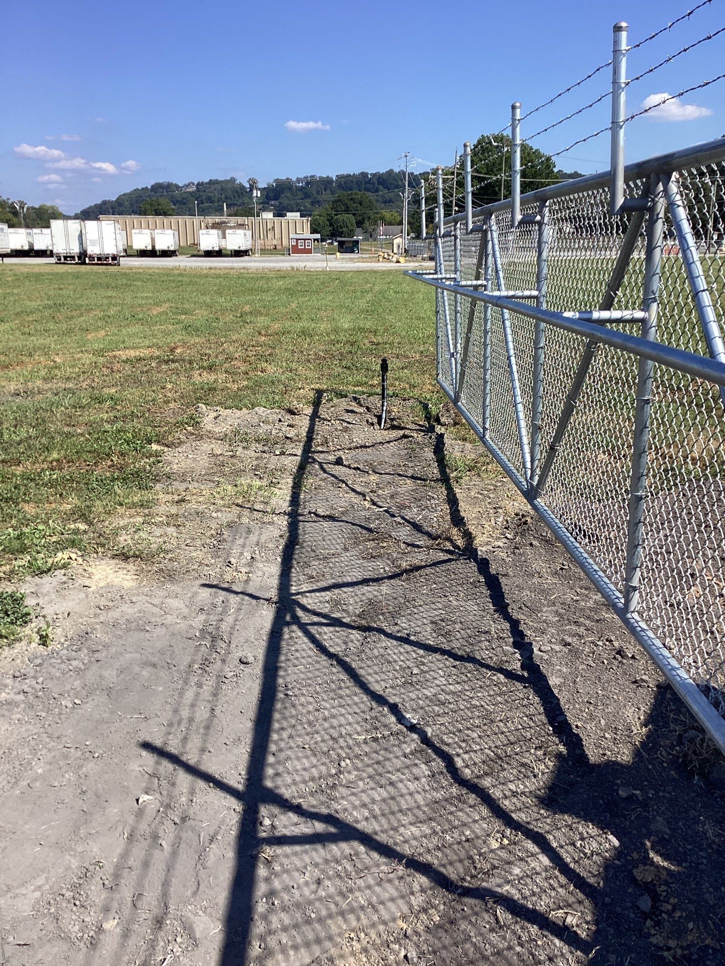 A chain-link fence borders a dirt path, casting a shadow on the ground, with grass and buildings in the background.