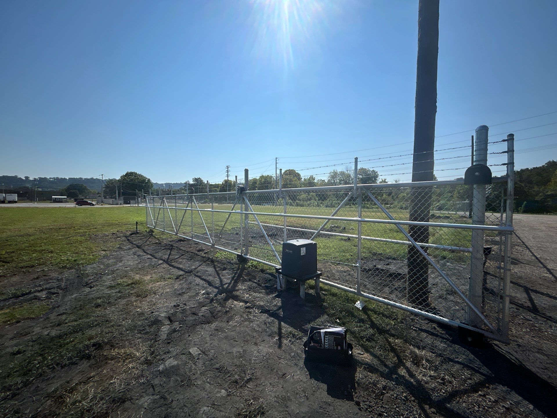 Metal security gate in front of a chain-link fence, under a bright blue sky. A grassy field is beyond.