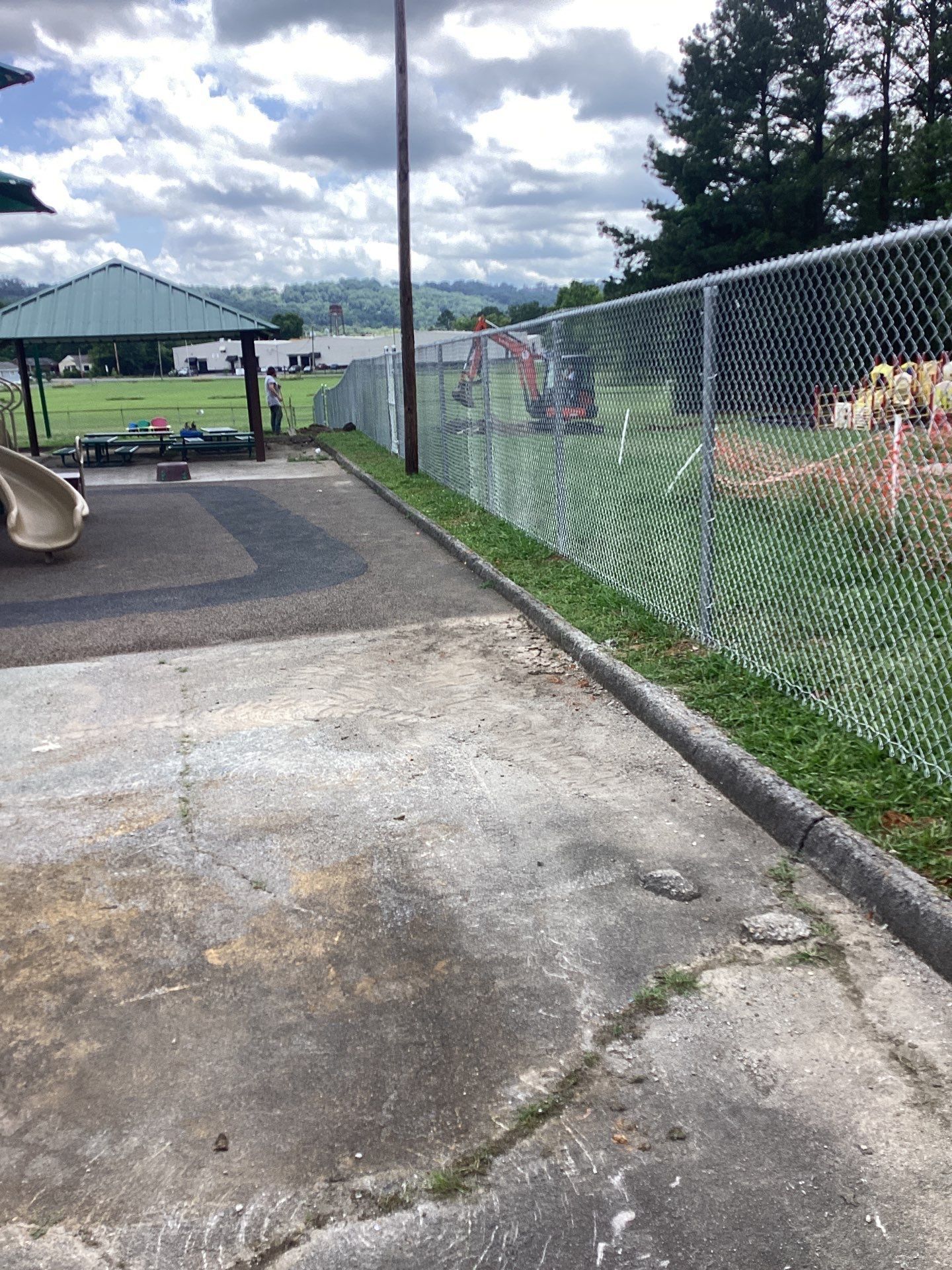 Concrete playground next to chain-link fence, with grass border and distant structures under a cloudy sky.