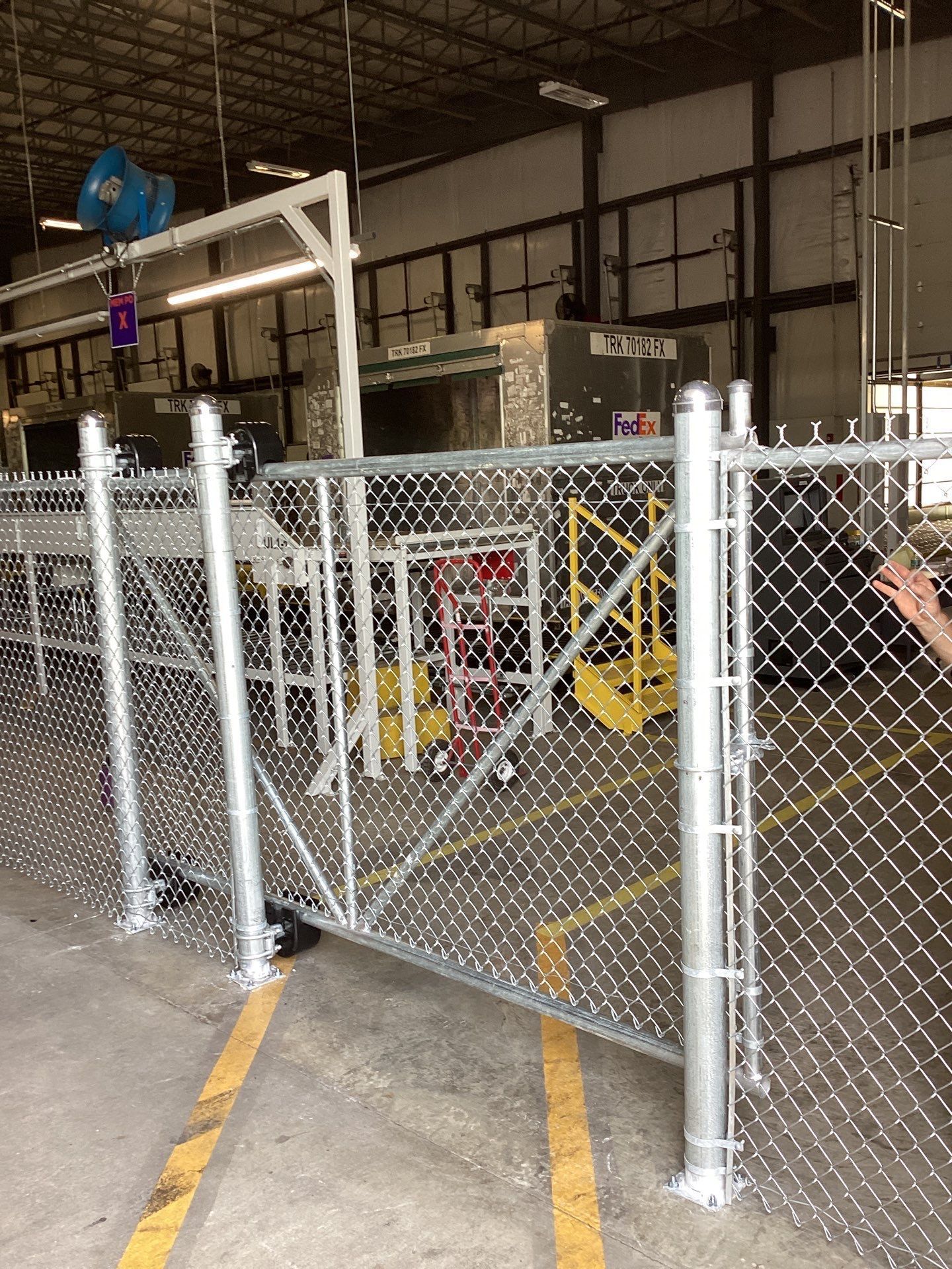 Chain-link fence with a gate, industrial setting. Yellow lines on floor, overhead equipment in background.