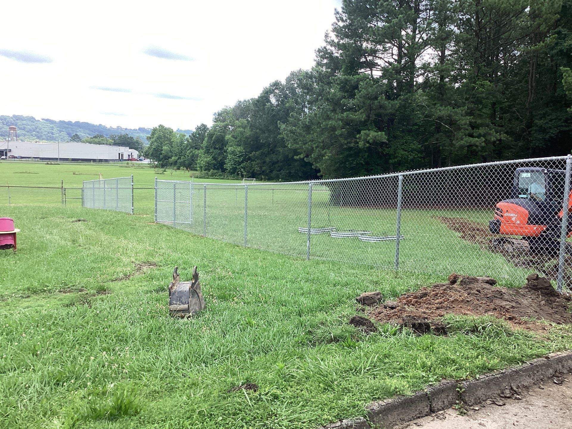 Chain-link fence enclosing a grassy area, small excavator, trees in the background.