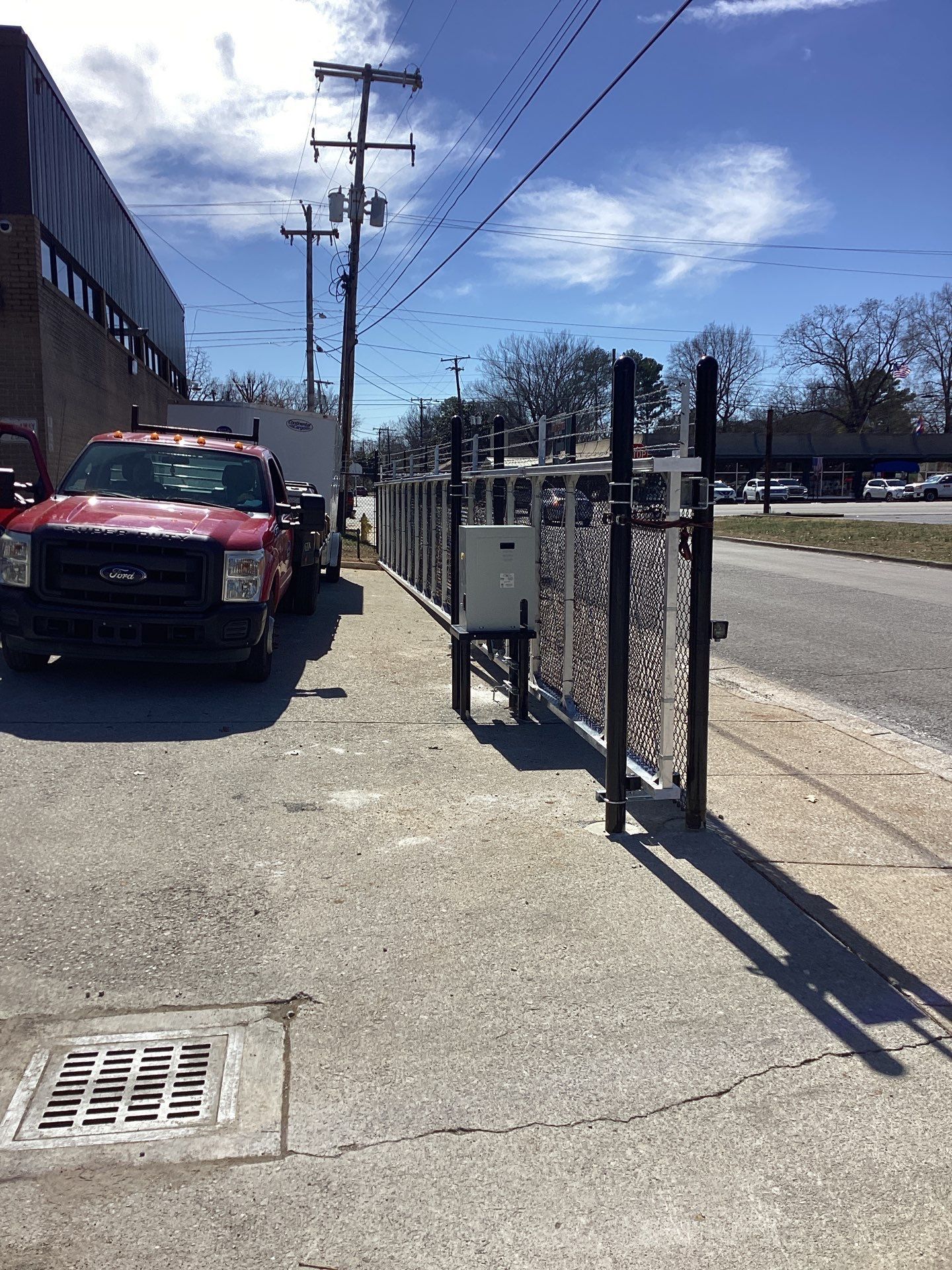 Red truck parked beside a black and silver fence on a concrete sidewalk, with power lines and a sunny sky.