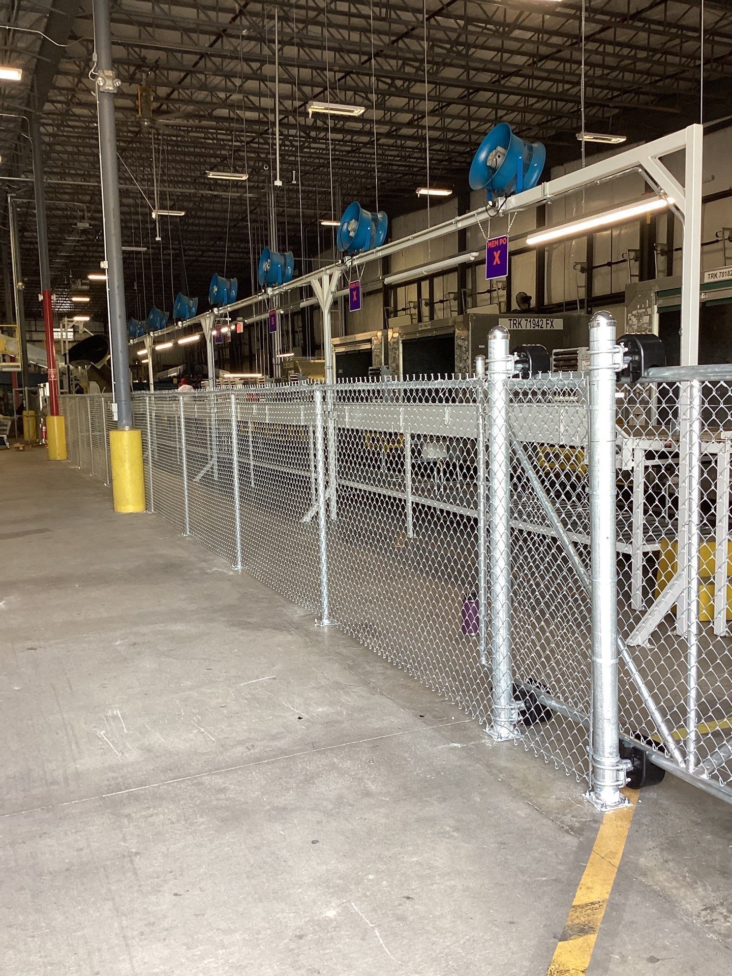 Chain-link fence in industrial setting, with overhead machinery, including blue fans.