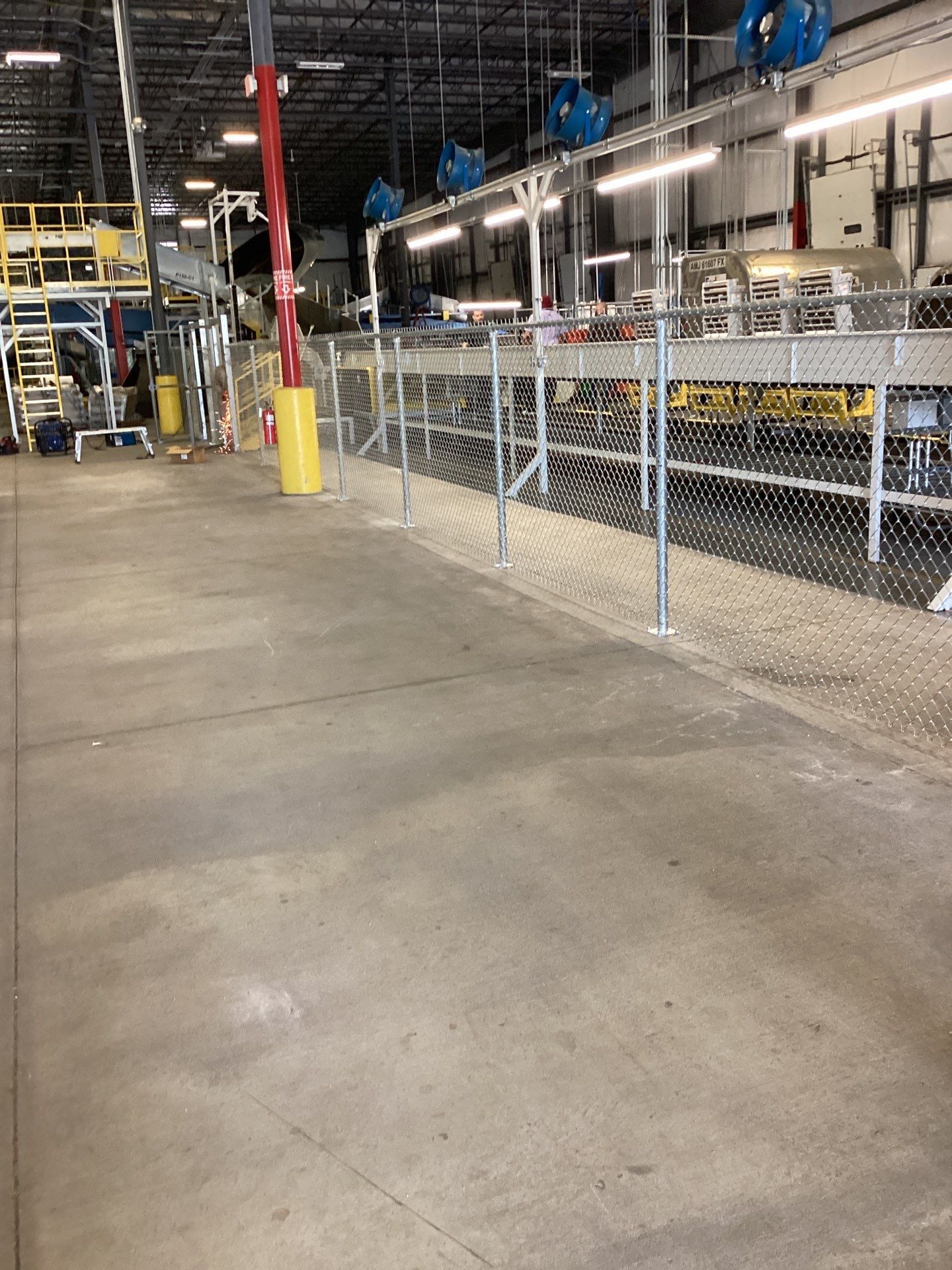 Warehouse interior with chain-link fence, concrete floor, overhead machinery, and various equipment.