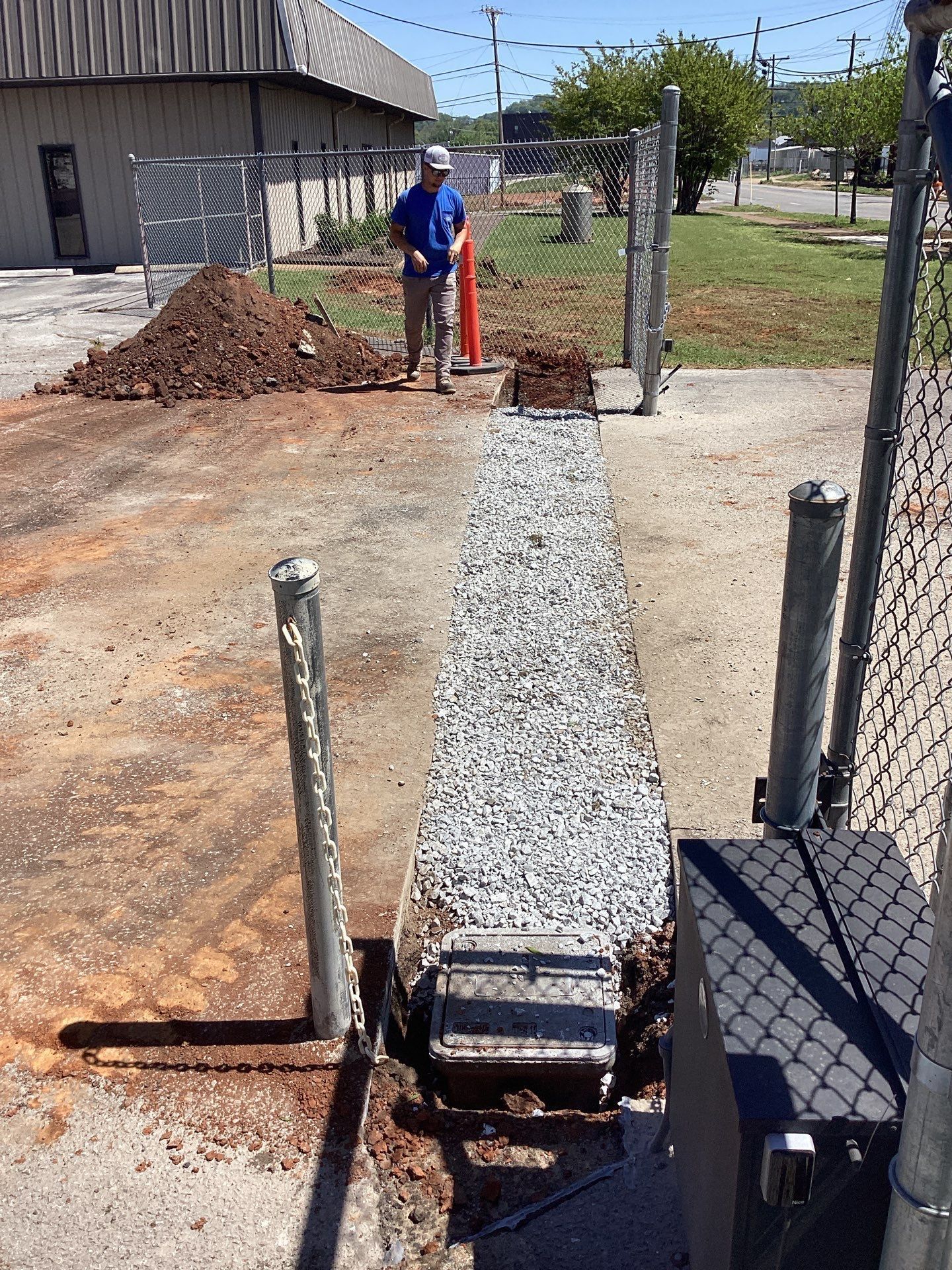 Man standing near gate with gravel path and dirt pile. Metal fence in industrial area.