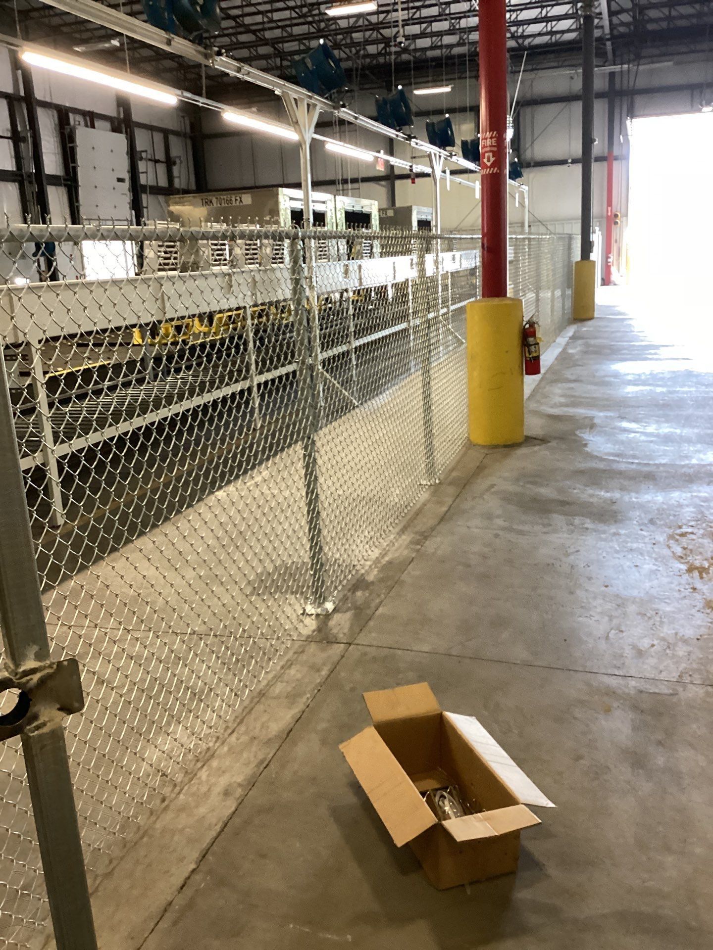 Inside a warehouse, a chain link fence lines a conveyor belt; an open cardboard box sits on the concrete floor.