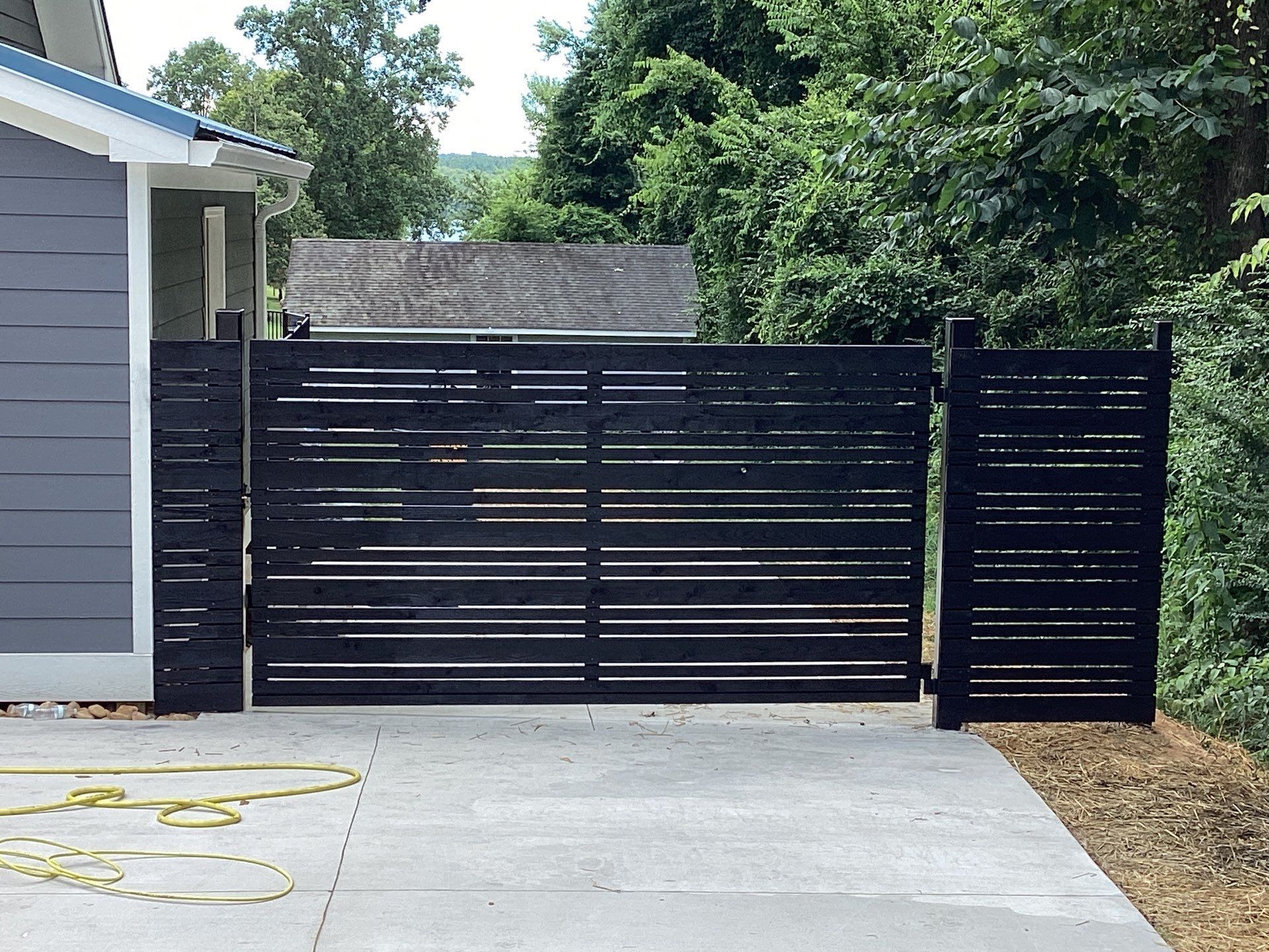Black horizontal slat gate and fence on a concrete driveway, next to a blue house.