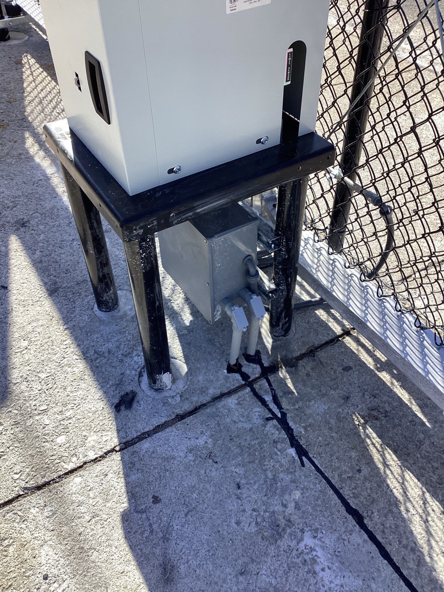 Gray electrical box on a metal stand next to a chain link fence.