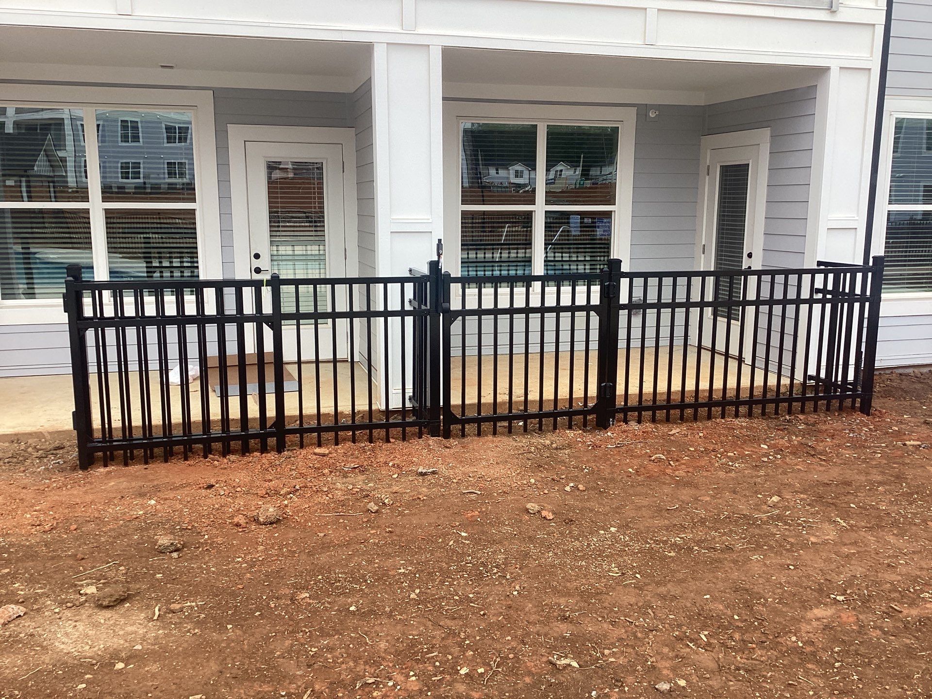 Black metal fence in front of a light gray building with glass doors and windows. The ground is dirt.
