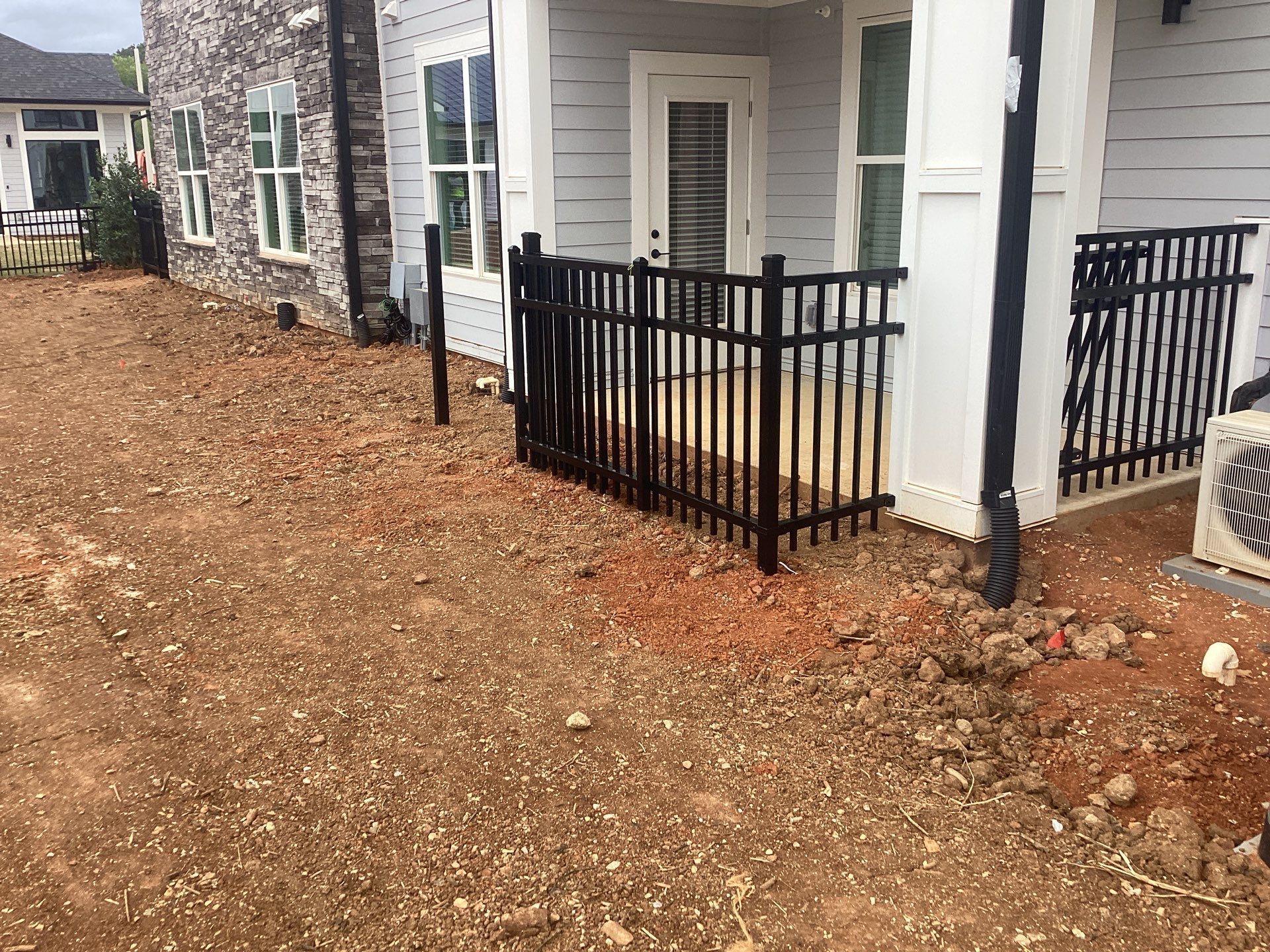 Black metal fence in front of a light gray building with a door, on a dirt patch.