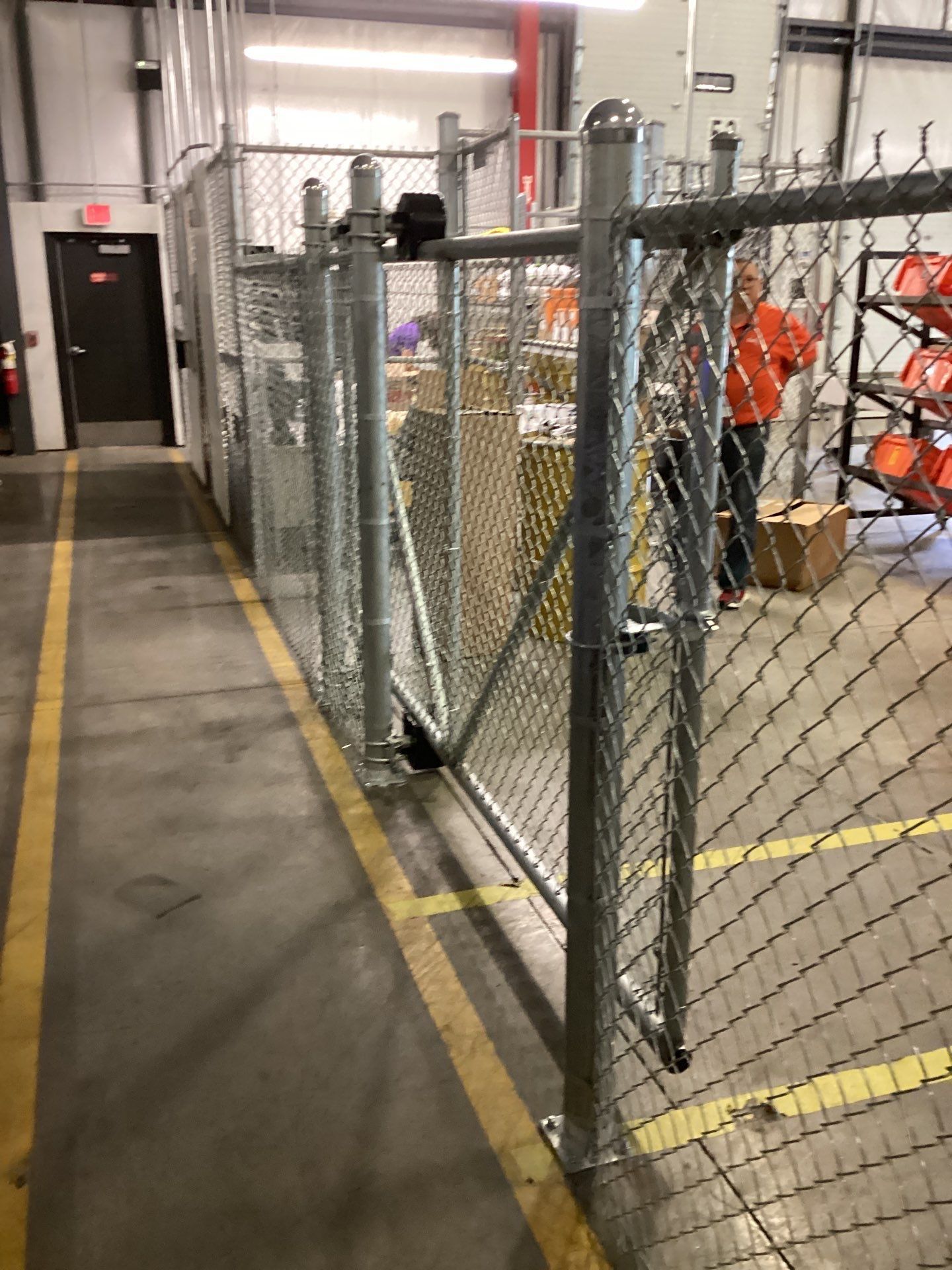 Metal chain link fence with a gate in a warehouse hallway, with yellow lines on the floor.