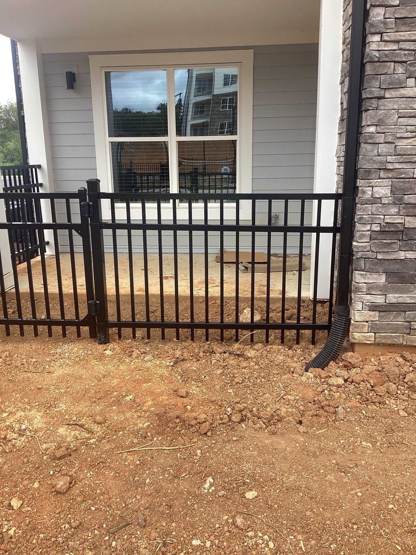 Black metal fence in front of a patio with a window, next to a stone wall and dirt ground.