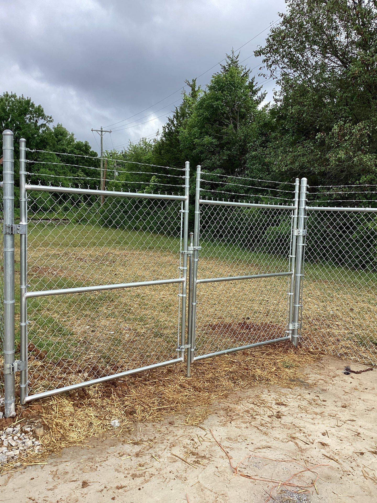 Chain link fence with double gates, topped with barbed wire, set against a grassy area with trees and overcast sky.
