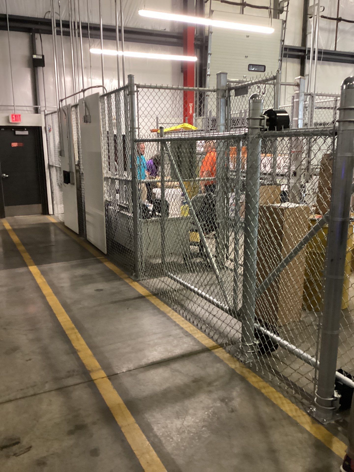 Chain-link fence encloses machinery in a warehouse. Yellow line on floor.