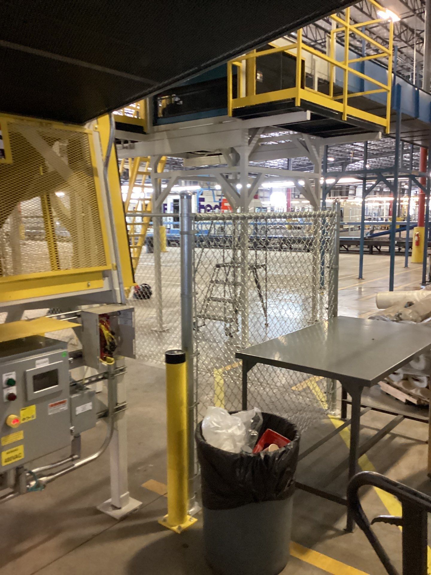 Industrial facility with conveyor system and safety fencing. Includes a trash can, control panel, and elevated platform.