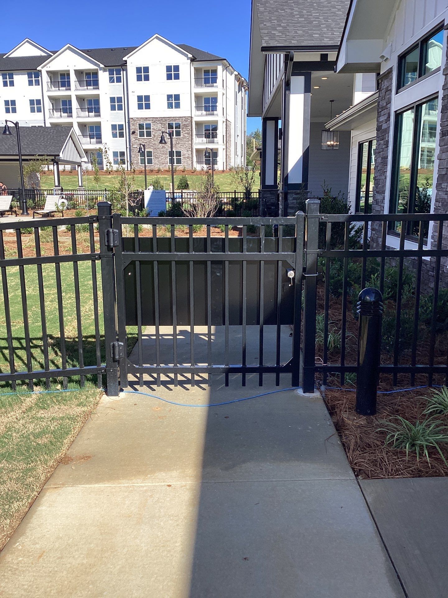 Black metal gate in a sidewalk, leading to a fenced area. Apartment building in background.