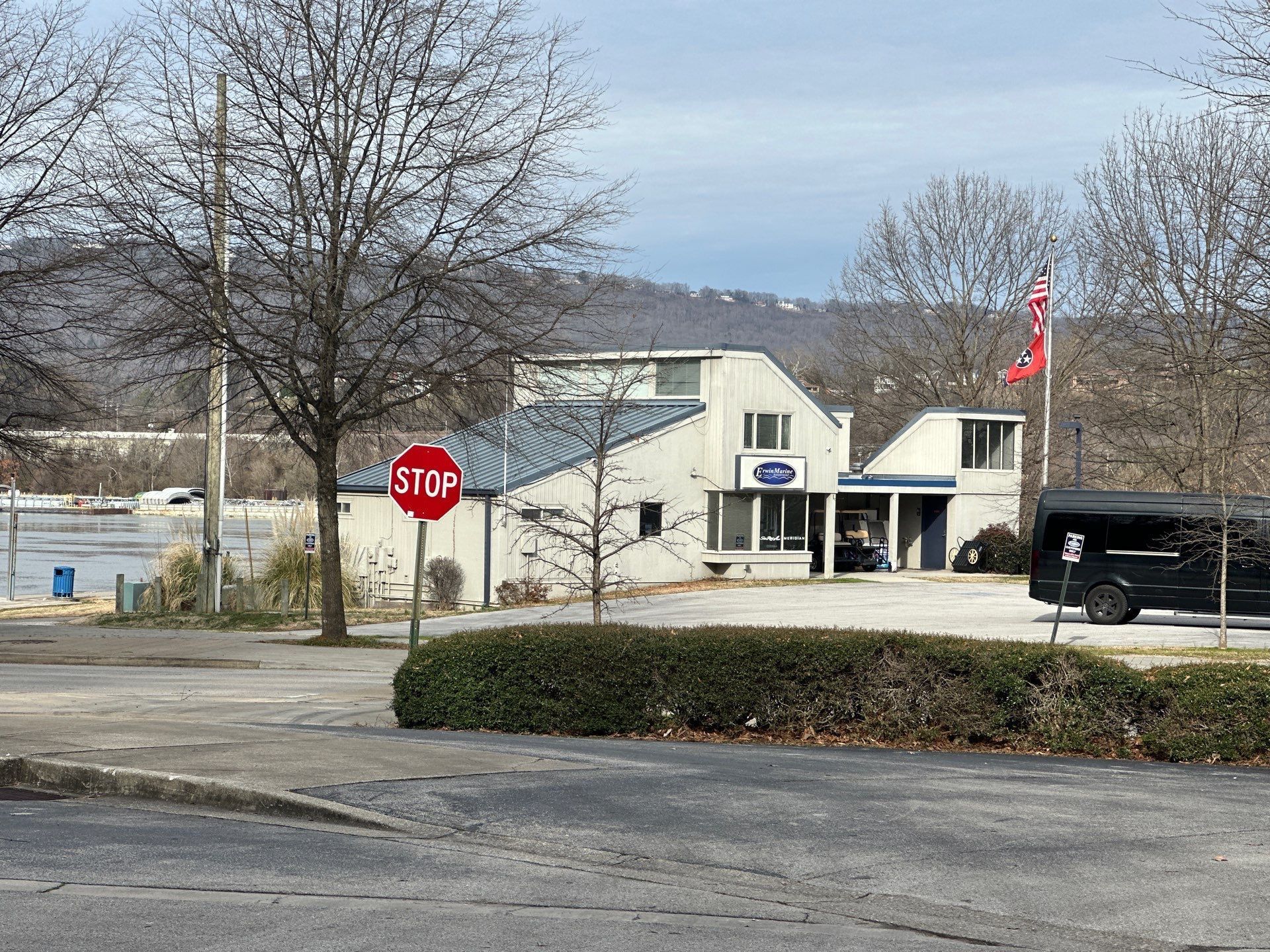 Building with stop sign in front, small bushes, a flag, and a van, with a body of water in the background.