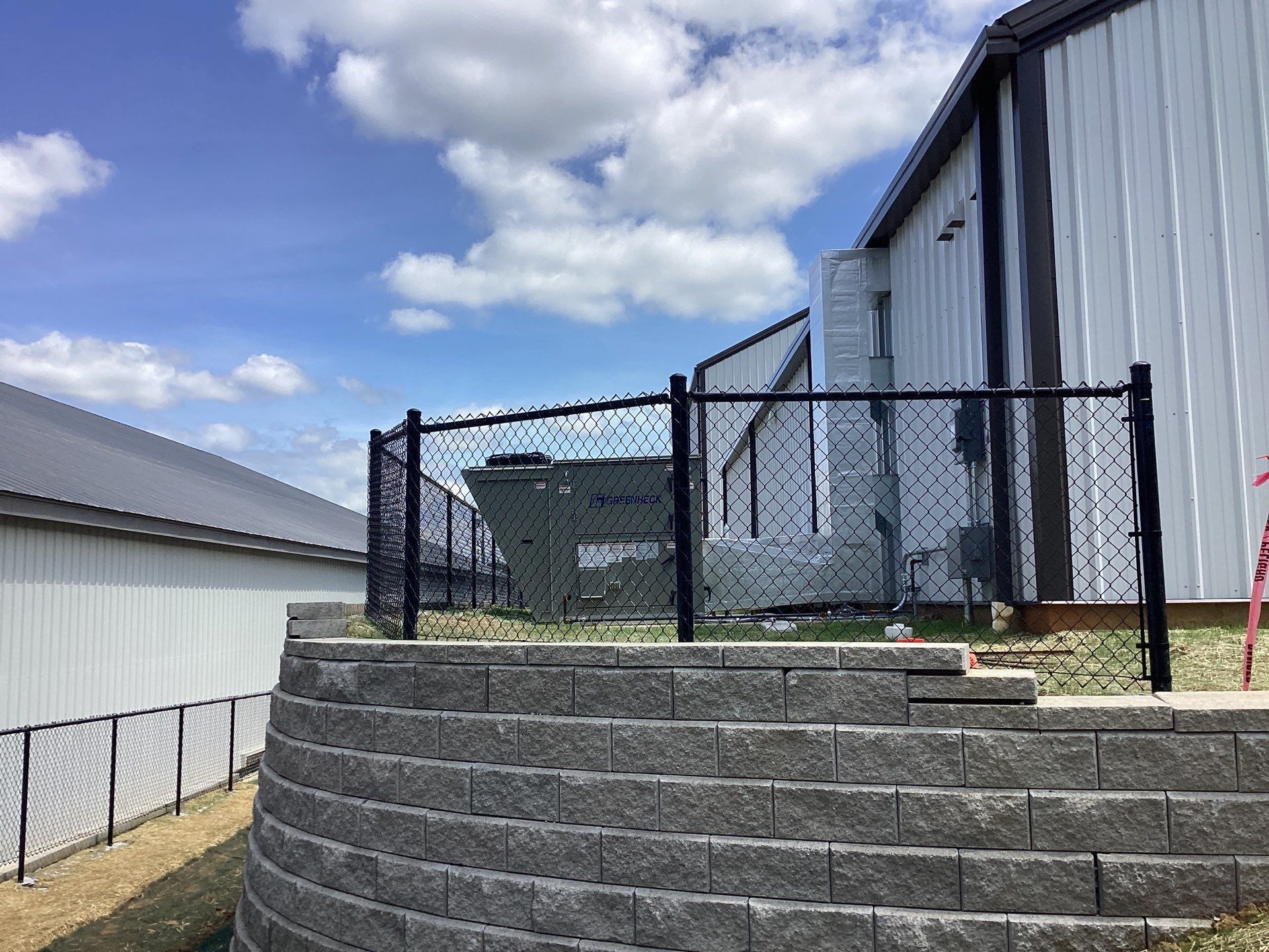 Black chain-link fence surrounds equipment next to a building on a gray stone retaining wall under a blue sky.