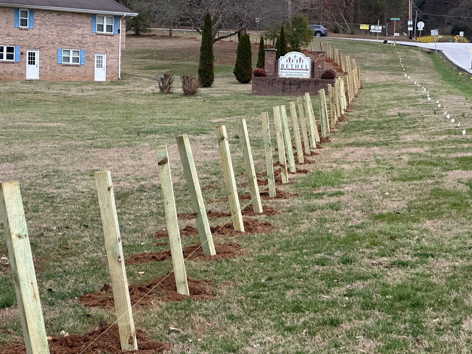 Wooden fence posts installed on a grassy hillside, with a building and sign in the background.