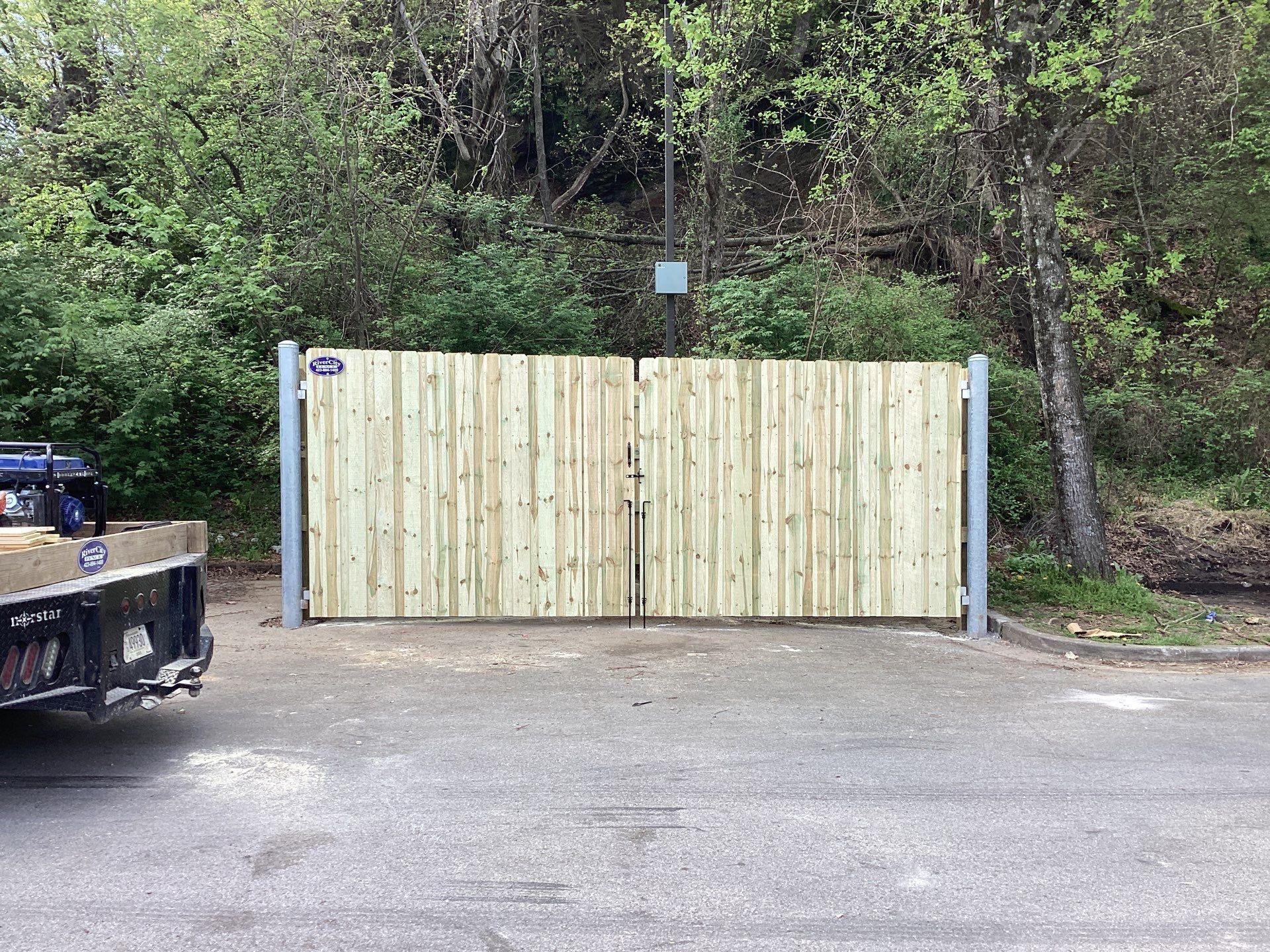 Wooden double gate, light wood color, set between gray metal posts, asphalt in front, trees behind.