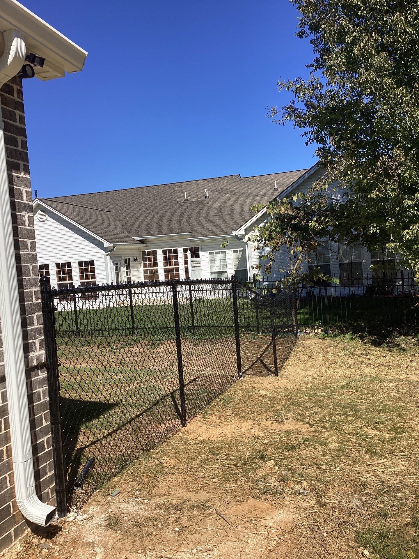 Black chain-link fence in a yard next to a white house with a brown roof and a blue sky.