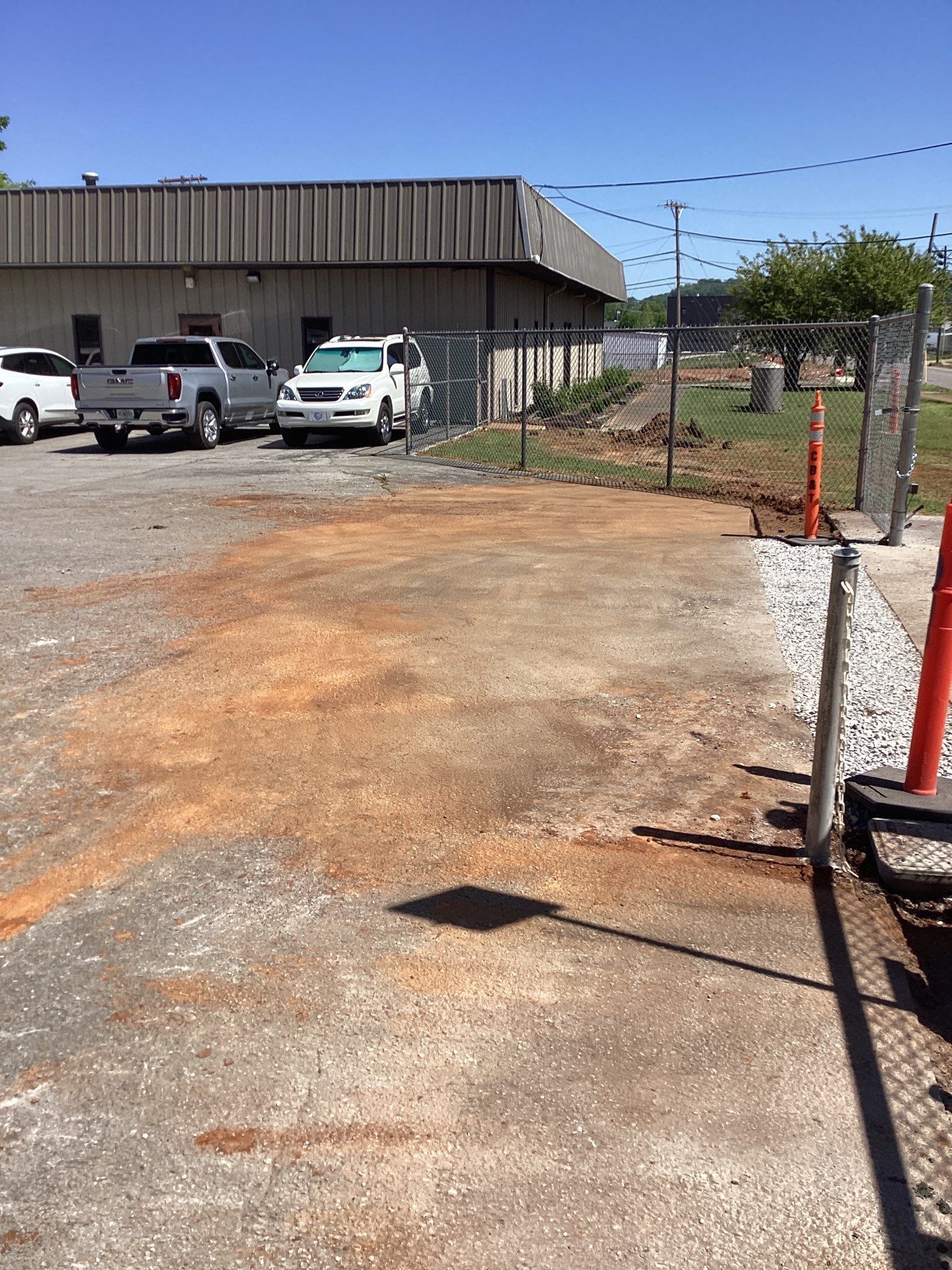 Paved lot with vehicles, building in the background. Open gate, orange cones. Sunny day, shadows.