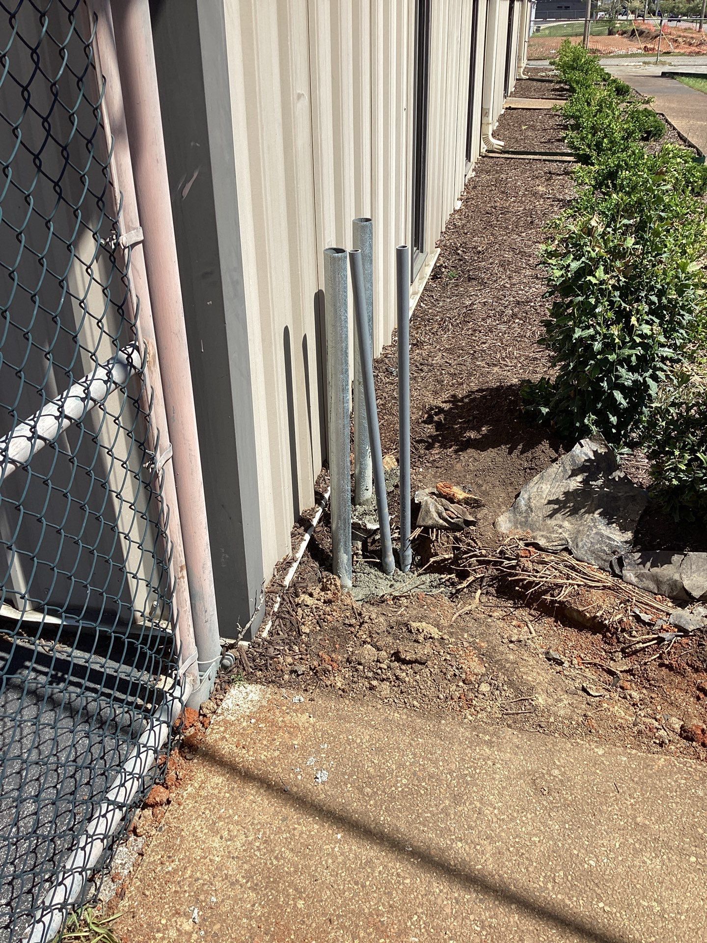 Metal pipes emerge from concrete near a building's wall, with a chain link fence in view.