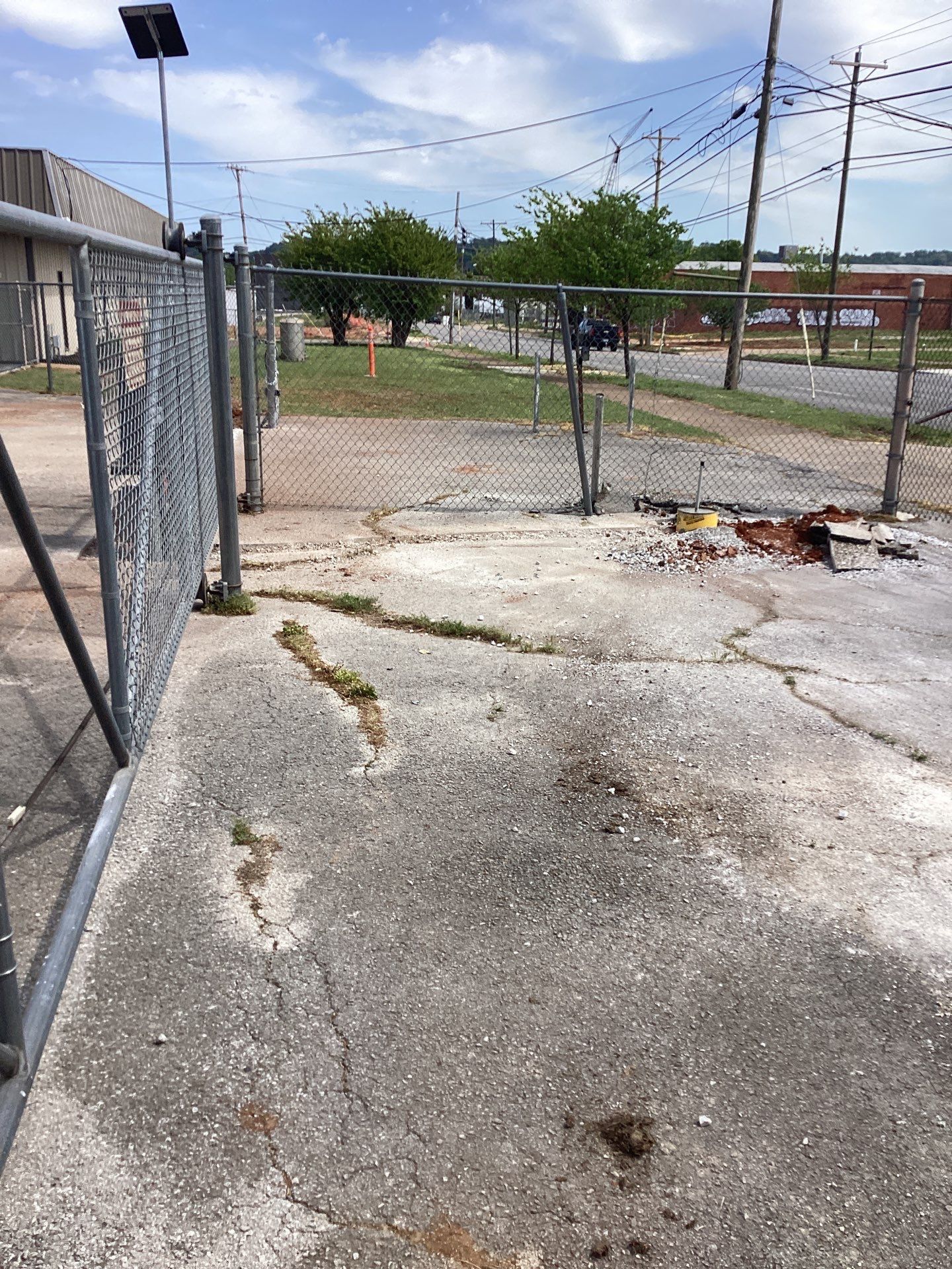Open chain-link gate on cracked concrete; grassy area beyond. Overhead power lines and buildings in background.