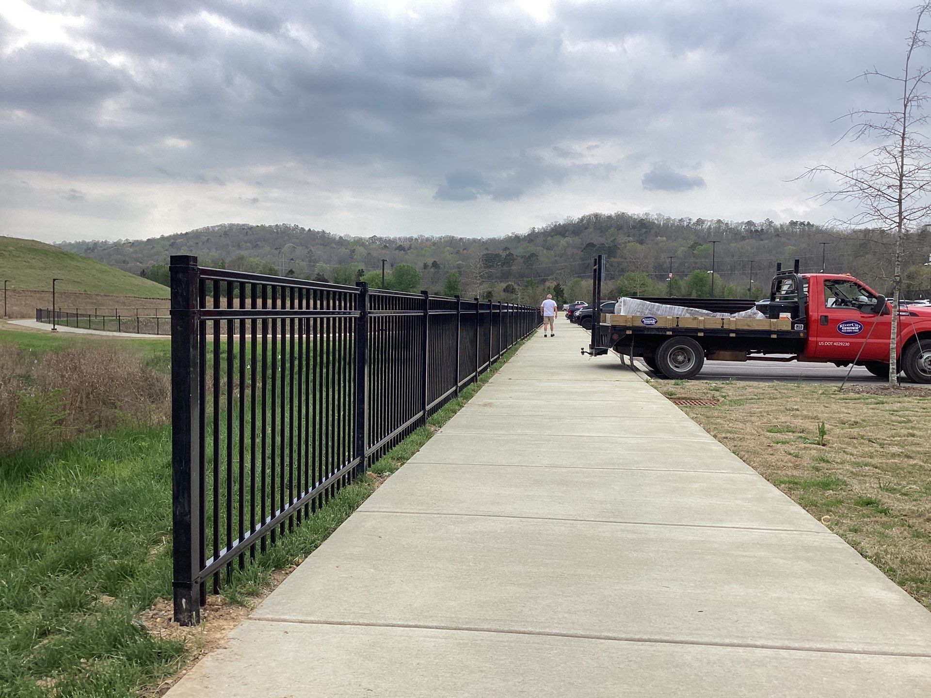 Black fence alongside a concrete path; red truck with supplies. Mountains in the distance under a cloudy sky.