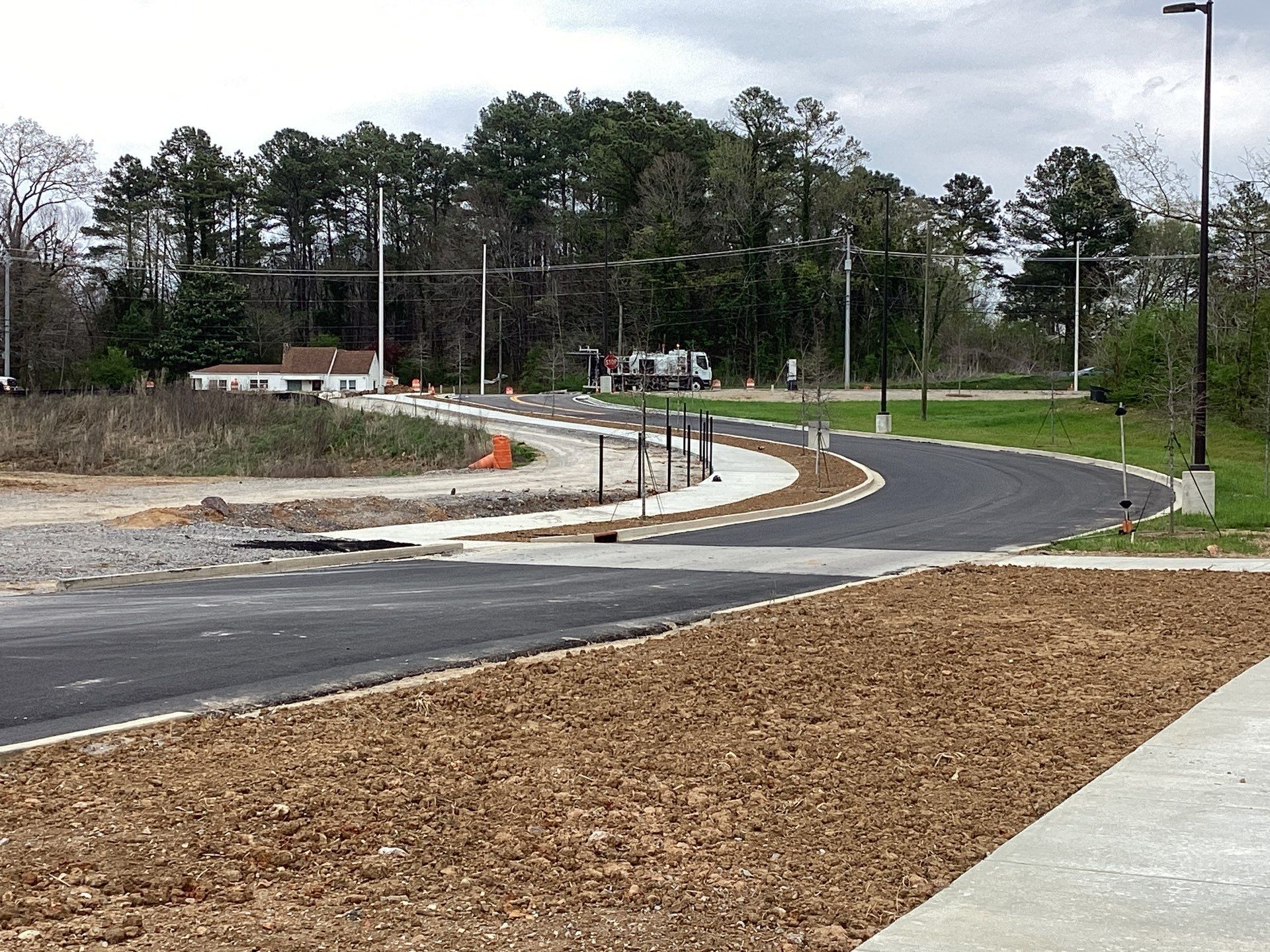 Newly paved road curving through a construction site, with dirt, trees, and streetlights.