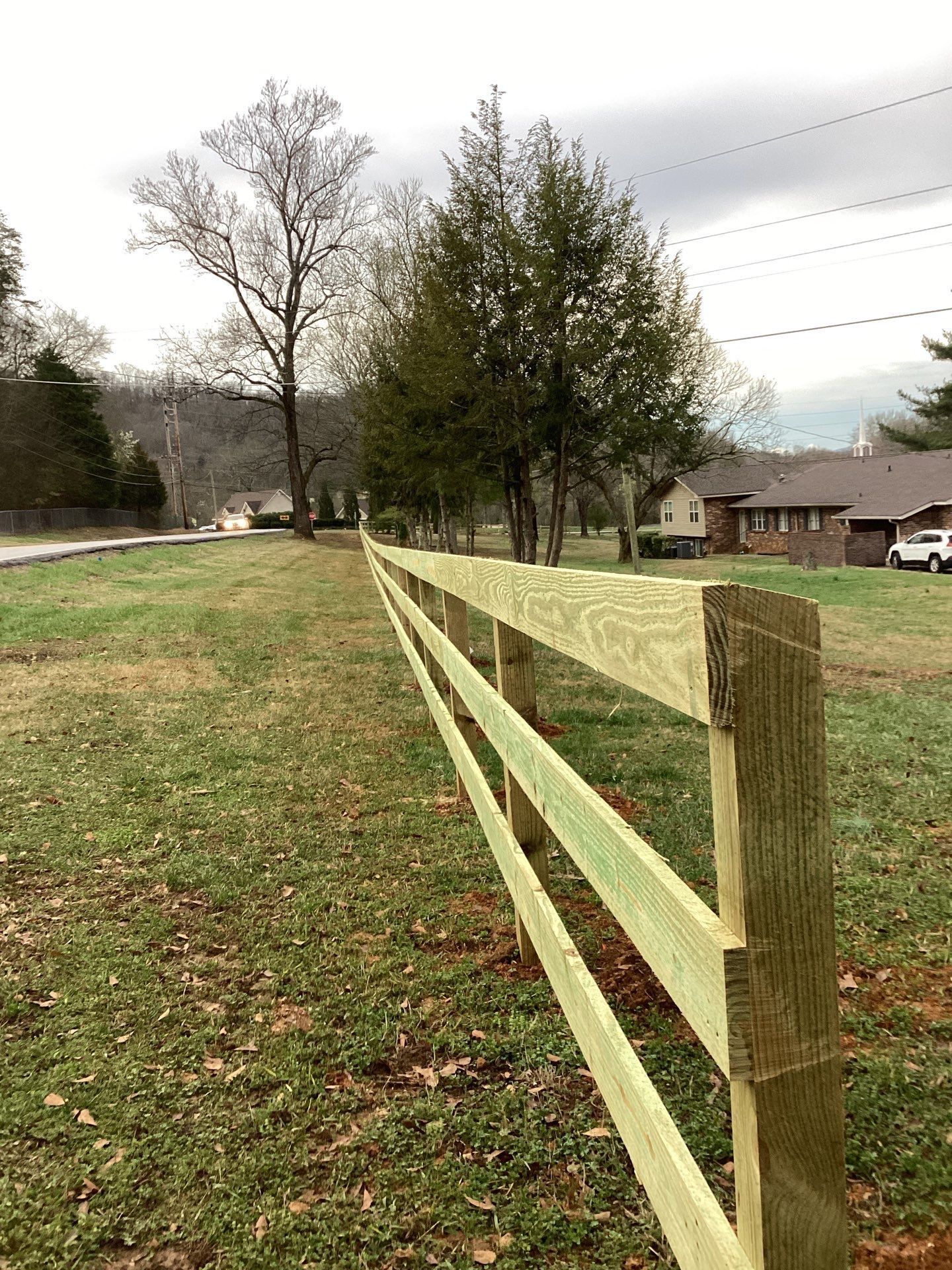 Wooden fence runs along grassy area with trees and houses in the background. Cloudy sky.