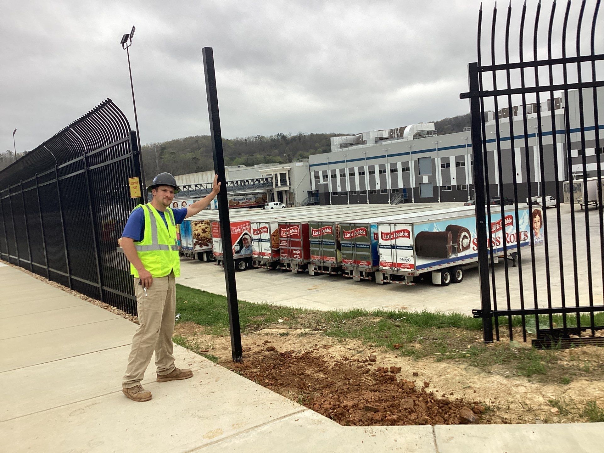 Man in work clothes near a fence, pointing at a pole. Trailers and a building are visible in the background.